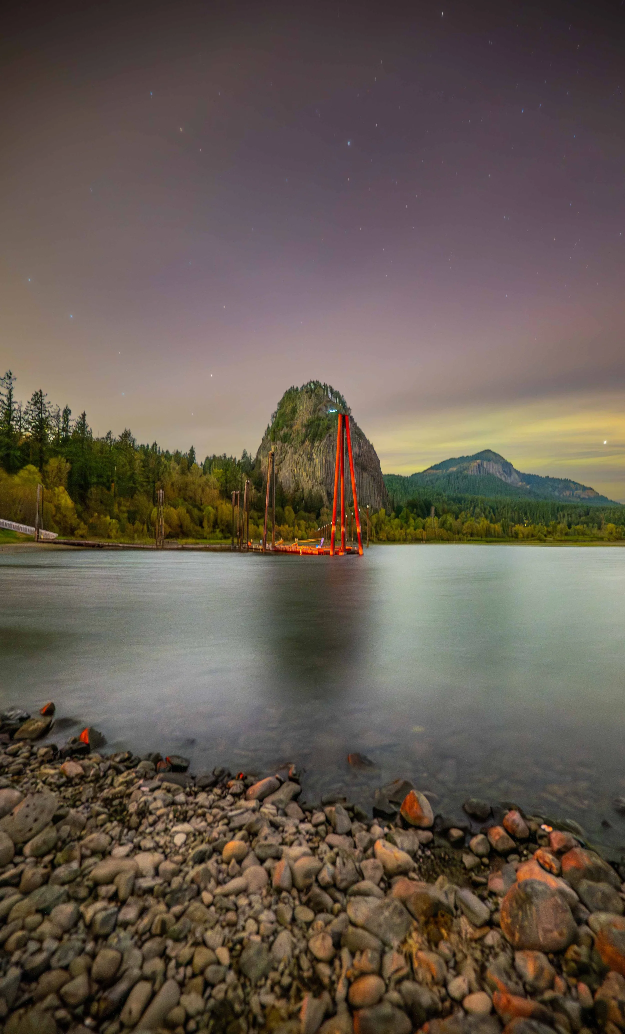 Nighttime view of a rocky shoreline with a large mountain in the background, a dam with red lights, and starry sky above.