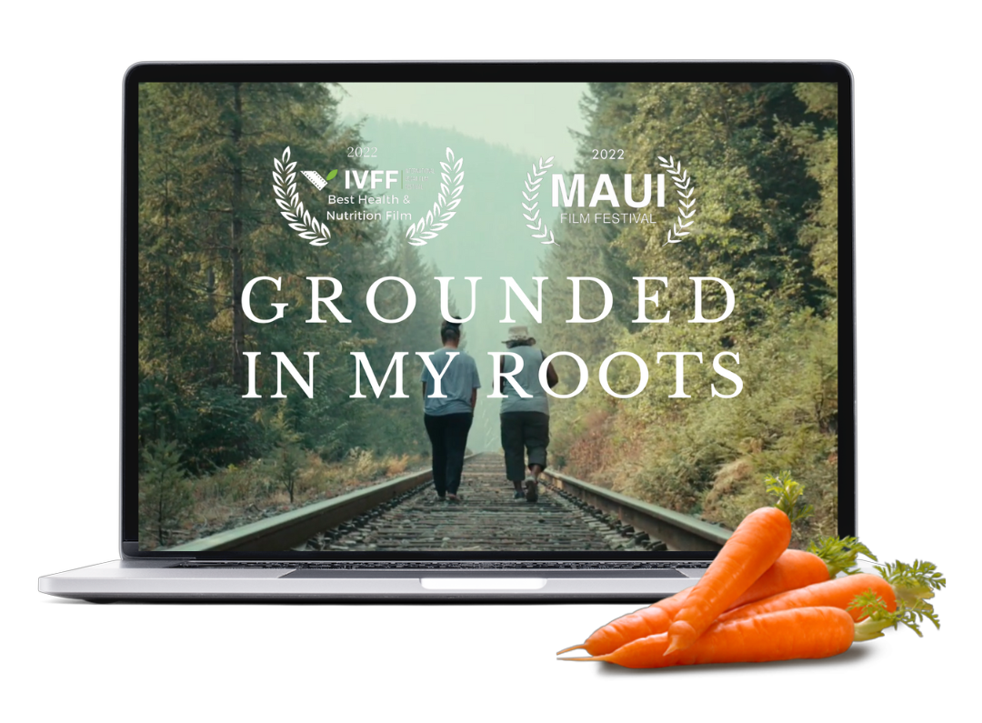 Laptop screen displaying a forest scene with two people walking along train tracks, with the text "Grounded in My Roots" and logos for IVFF and Maui Film Festival; carrots are placed in front of the laptop.