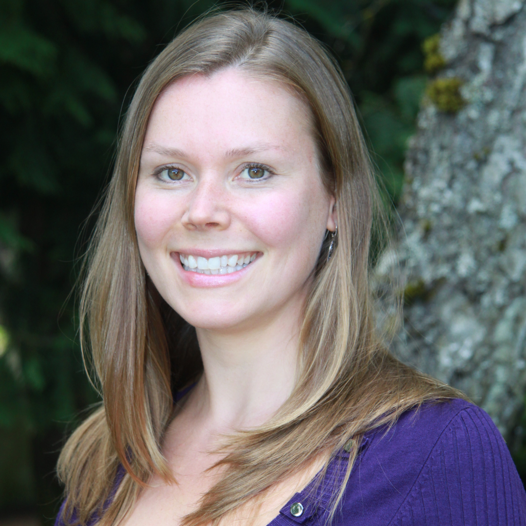 A smiling woman with long, straight, light brown hair, wearing a purple top, stands outdoors near a tree with green foliage in the background.