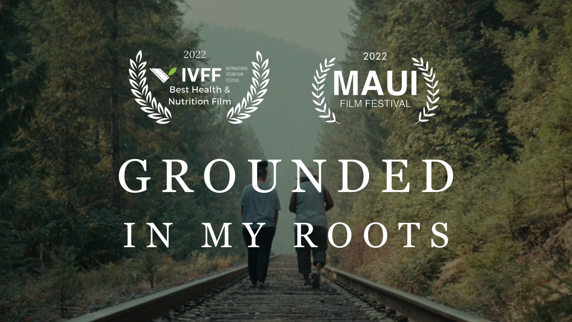 Two people walking on railway tracks through a forest with trees on either side, with text overlays indicating awards and festival information for a film titled 'Grounded in My Roots'.