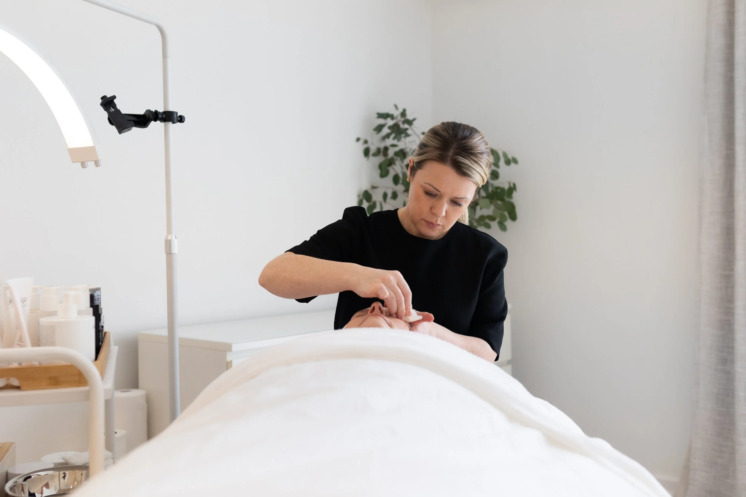A woman providing facial treatment to a person lying on a treatment bed in a spa or clinic room, with skincare products and equipment around. SKN Haus Balmain
