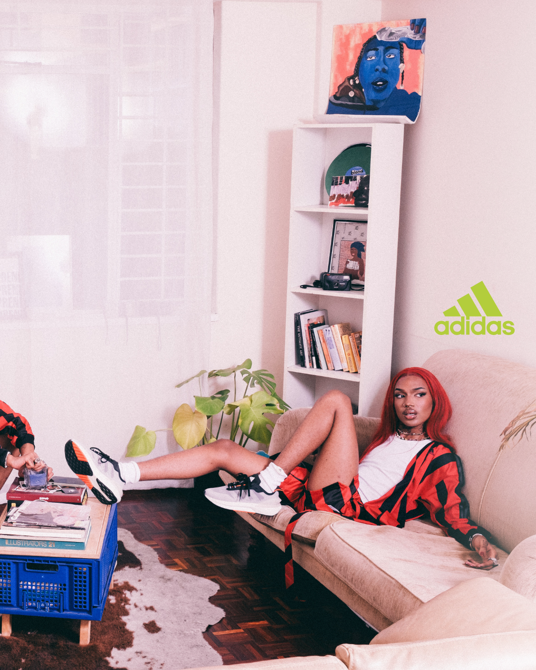 A woman with red hair relaxes on a beige sofa in a living room, wearing a white t-shirt, red and black checkered shirt, and black sneakers. The room has a white shelf with art, books, and decor, a green plant, and an Adidas logo on the wall.