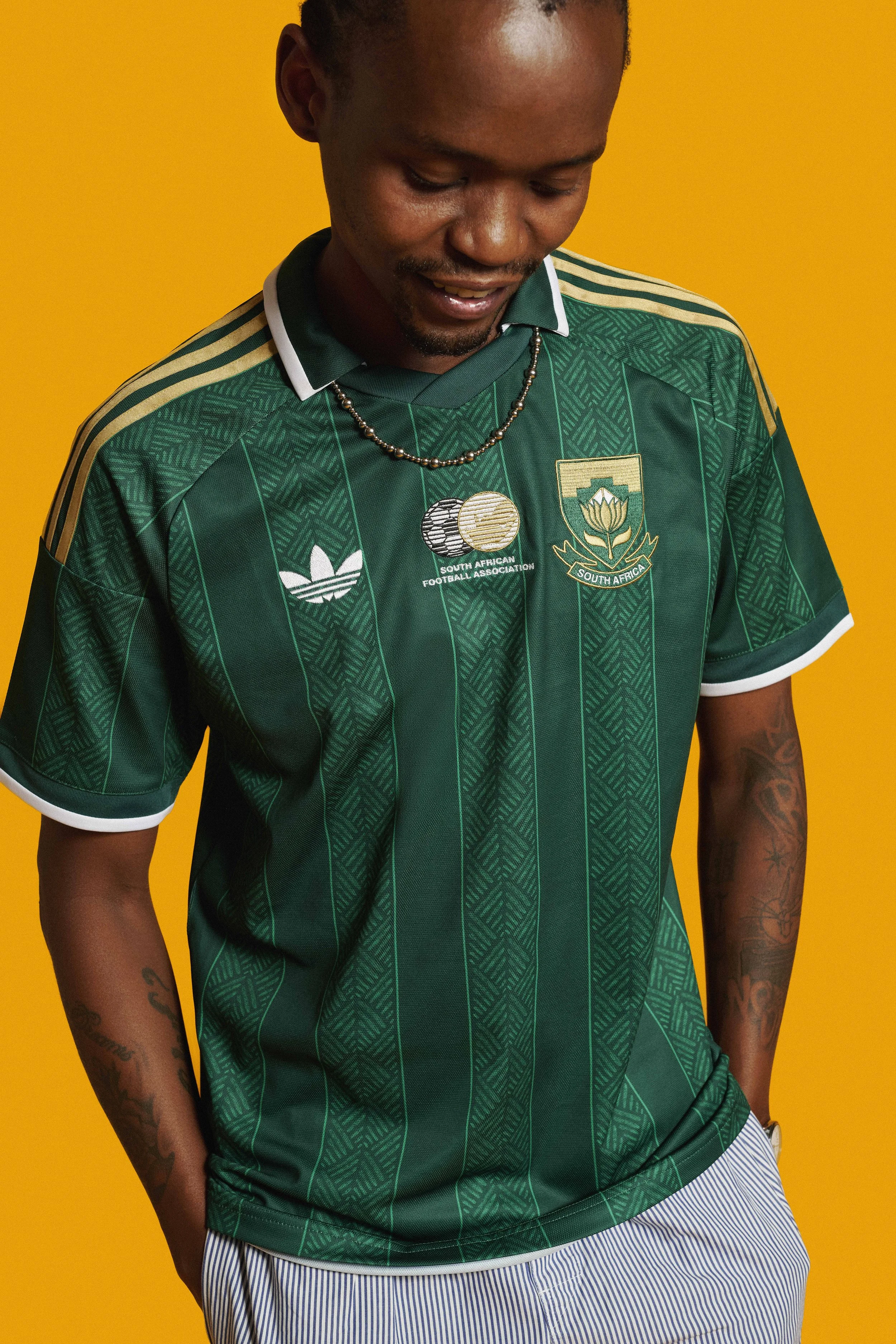 adidas Football safa world cup 2026 jersey launch Bafana Bafana athletes. Away jersey. A man wearing a green South African football jersey with gold and white details, standing against a yellow background, looking down with hands in pockets.