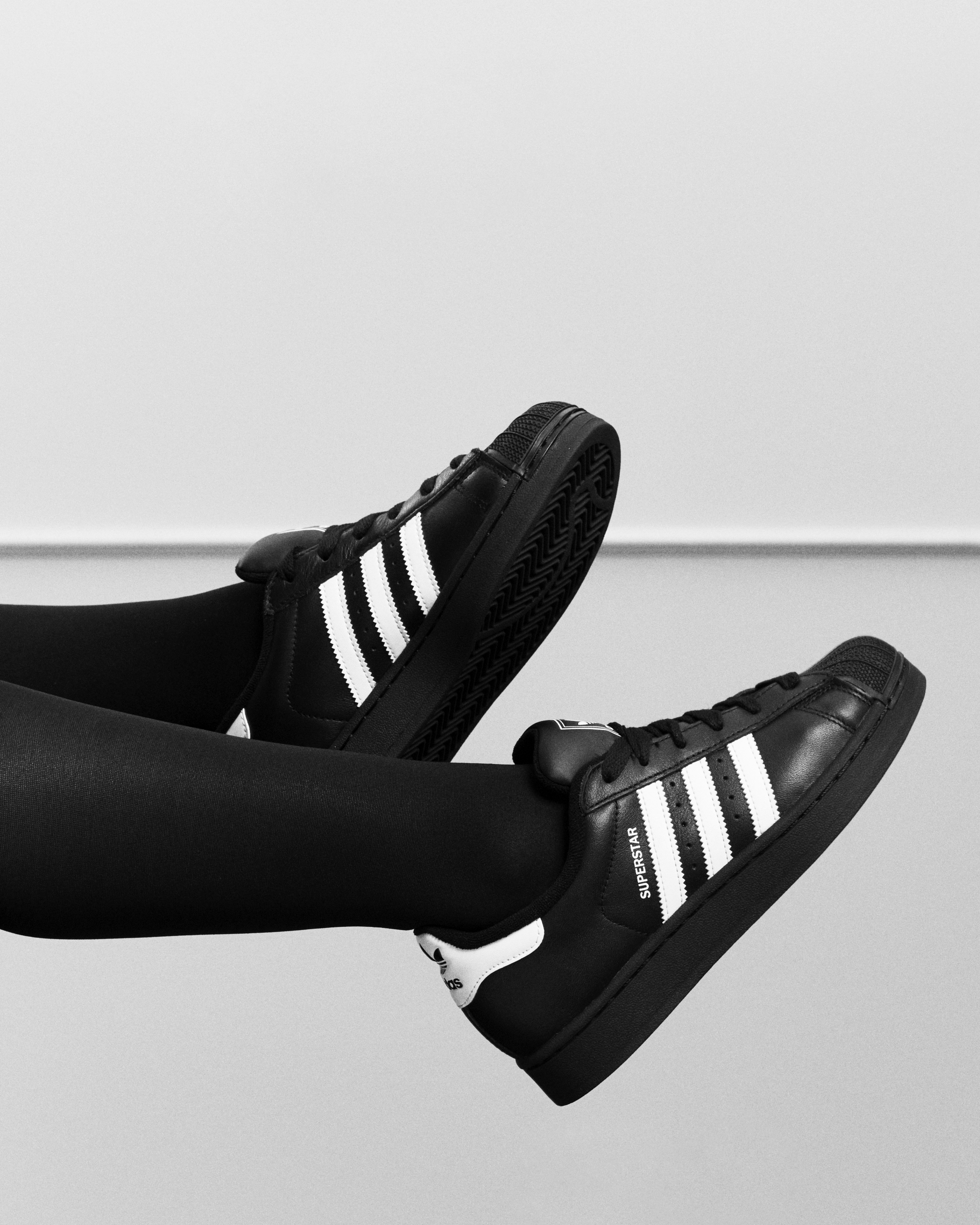 Uncle Waffles for adidas originals superstar spring summer 2026 campaign, wearing black leggings and black Adidas Superstar sneakers with white stripes, lifting foot in the air against a plain background.