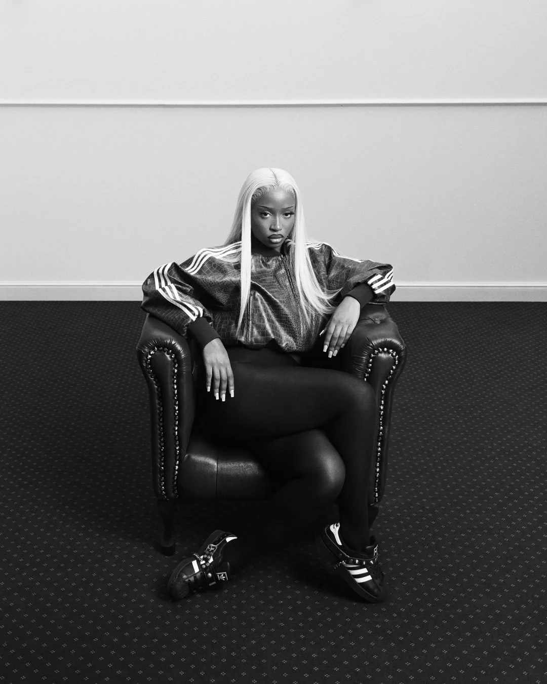 Uncle Waffles for adidas originals superstar spring summer 2026 campaign, wearing black leggings and black Adidas Superstar sneakers with white stripes, lifting foot in the air against a plain background.