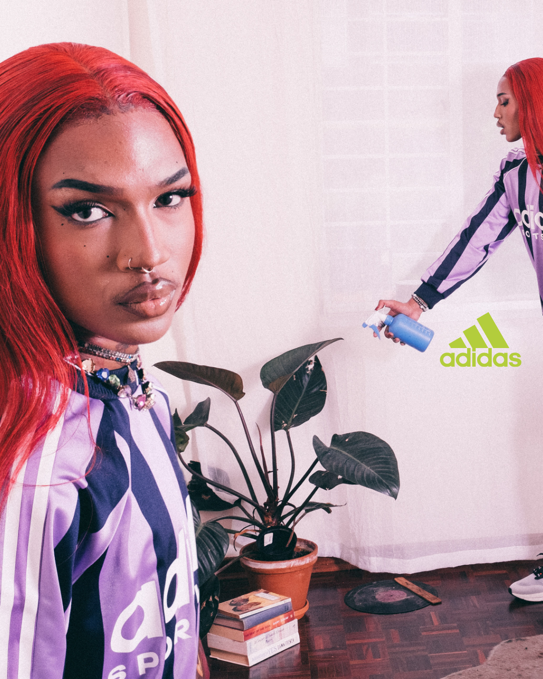 A woman with bright red hair and makeup, wearing a purple Adidas jersey, stands indoors next to a large potted plant and a stack of books. Another woman in a matching jersey is spraying disinfectant in the background, with the Adidas logo visible.