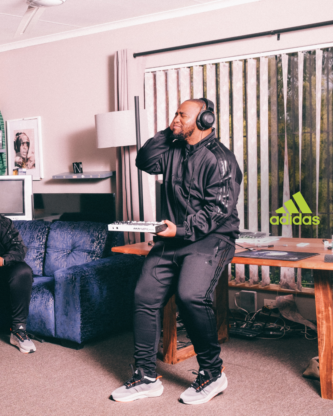 A man wearing a black Adidas tracksuit and headphones is holding a small keyboard and singing with closed eyes in a living room with a couch, a floor lamp, and a window with vertical blinds.