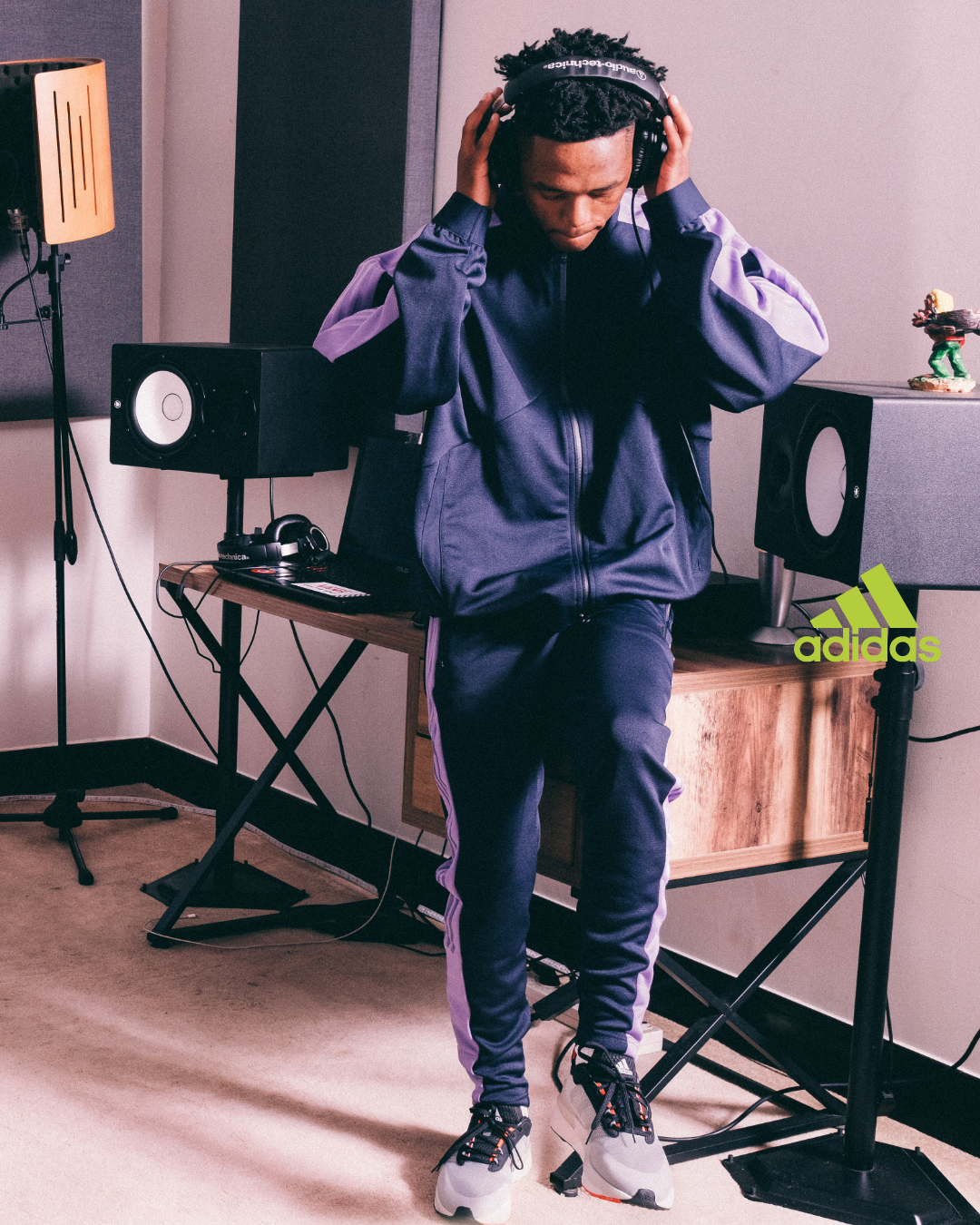 Young man in athletic clothing wearing headphones in a recording studio with speakers, a microphone stand, and an Adidas logo on the wall.