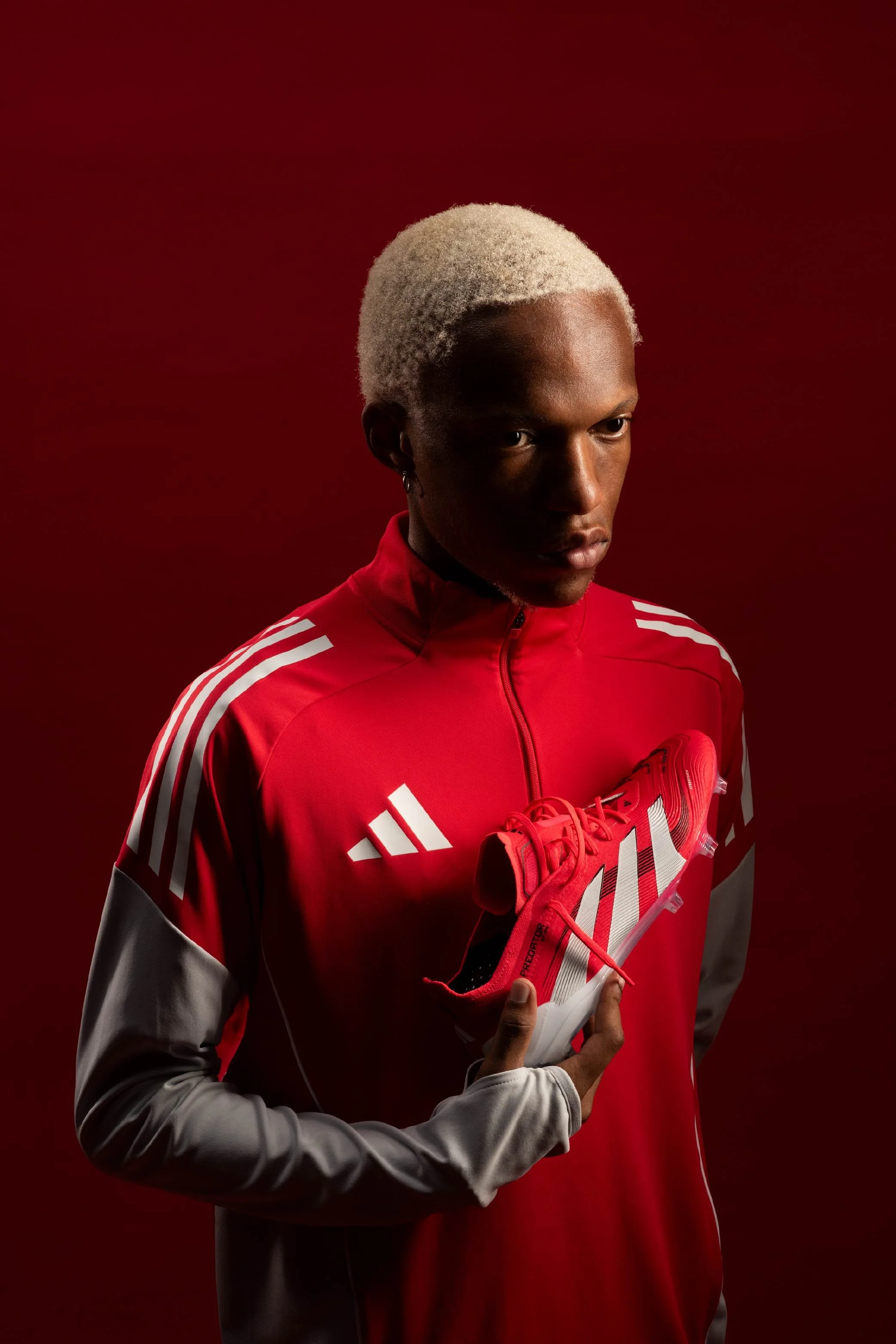 An athlete with platinum blonde hair in a red and grey Adidas sports jacket holding a red and white Adidas soccer cleat, standing against a dark red background.
