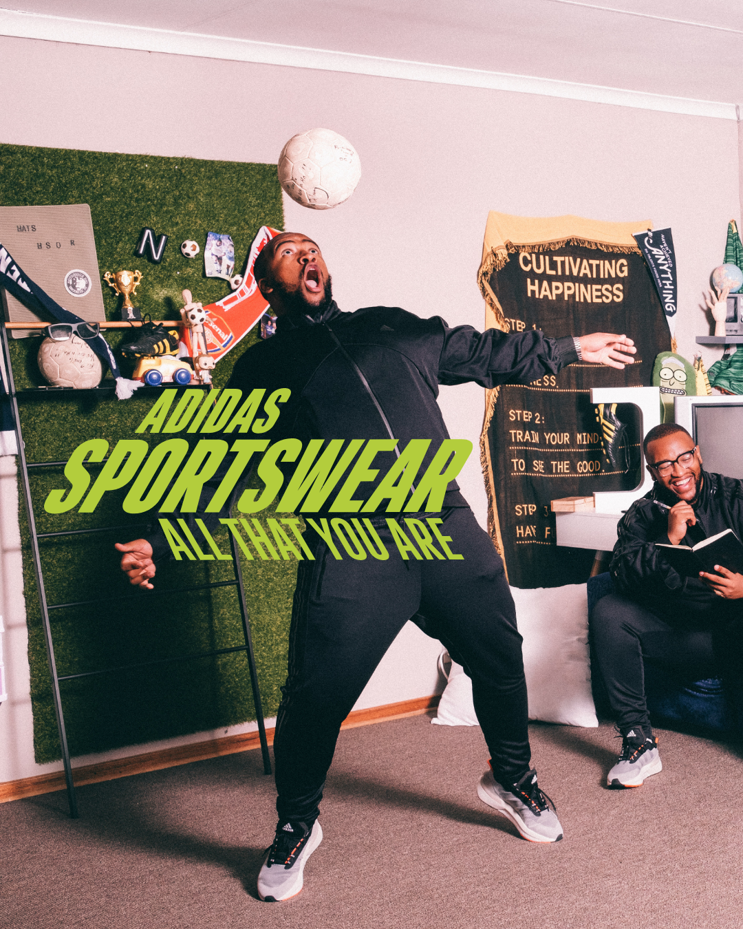 Two men in an indoor room with sports and motivational decor, one is actively kicking a white soccer ball while the other is sitting on a chair laughing. Room has sports memorabilia, a black banner with gold text, and a green grass wall panel. Overlaid text reads 'ADIDAS SPORTSWEAR ALL THAT YOU ARE.'