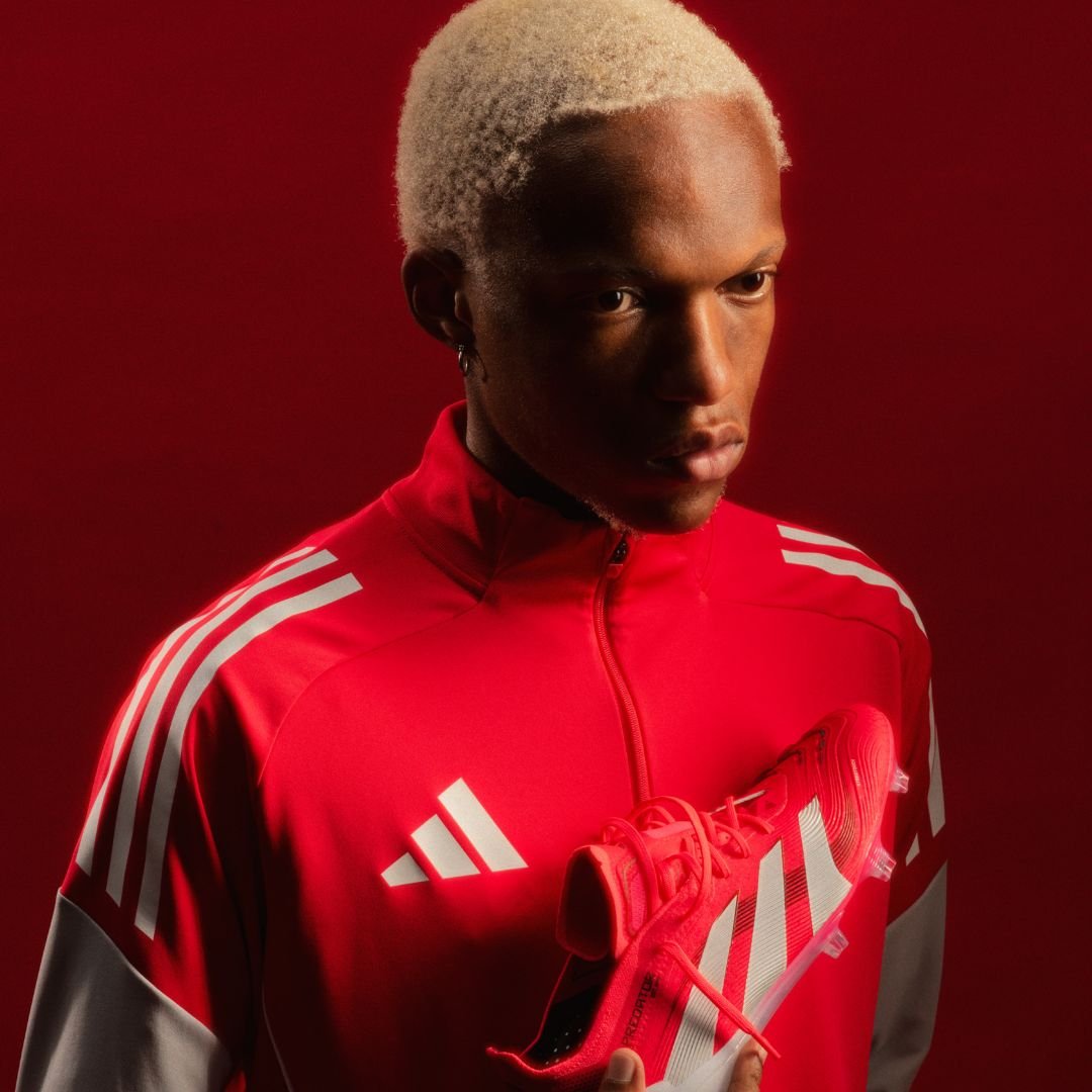 An athlete with platinum blonde hair in a red and grey Adidas sports jacket holding a red and white Adidas soccer cleat, standing against a dark red background.