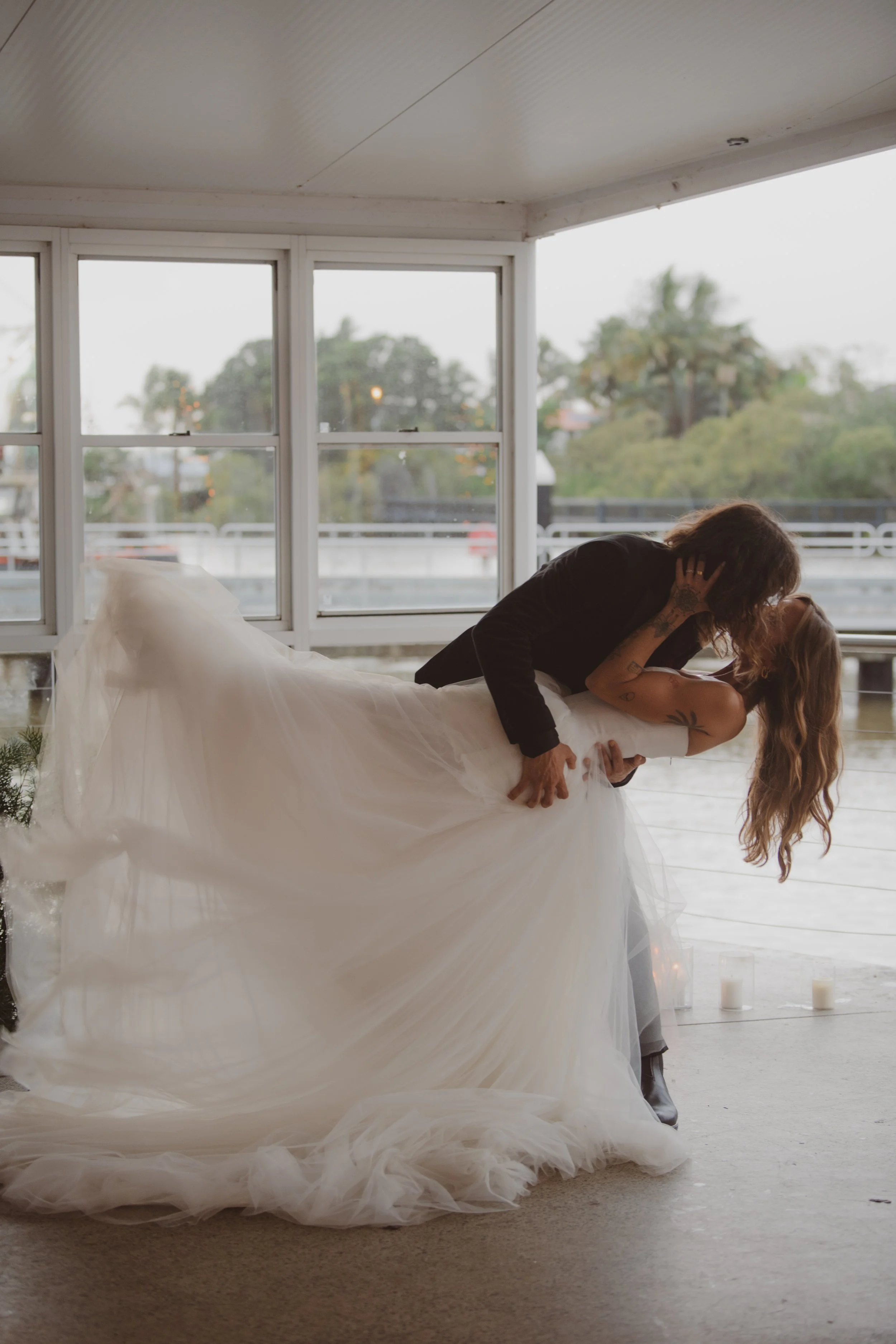 A couple shares a romantic dance in front of large windows with green trees outside, candles on the floor, and natural light filling the room.
