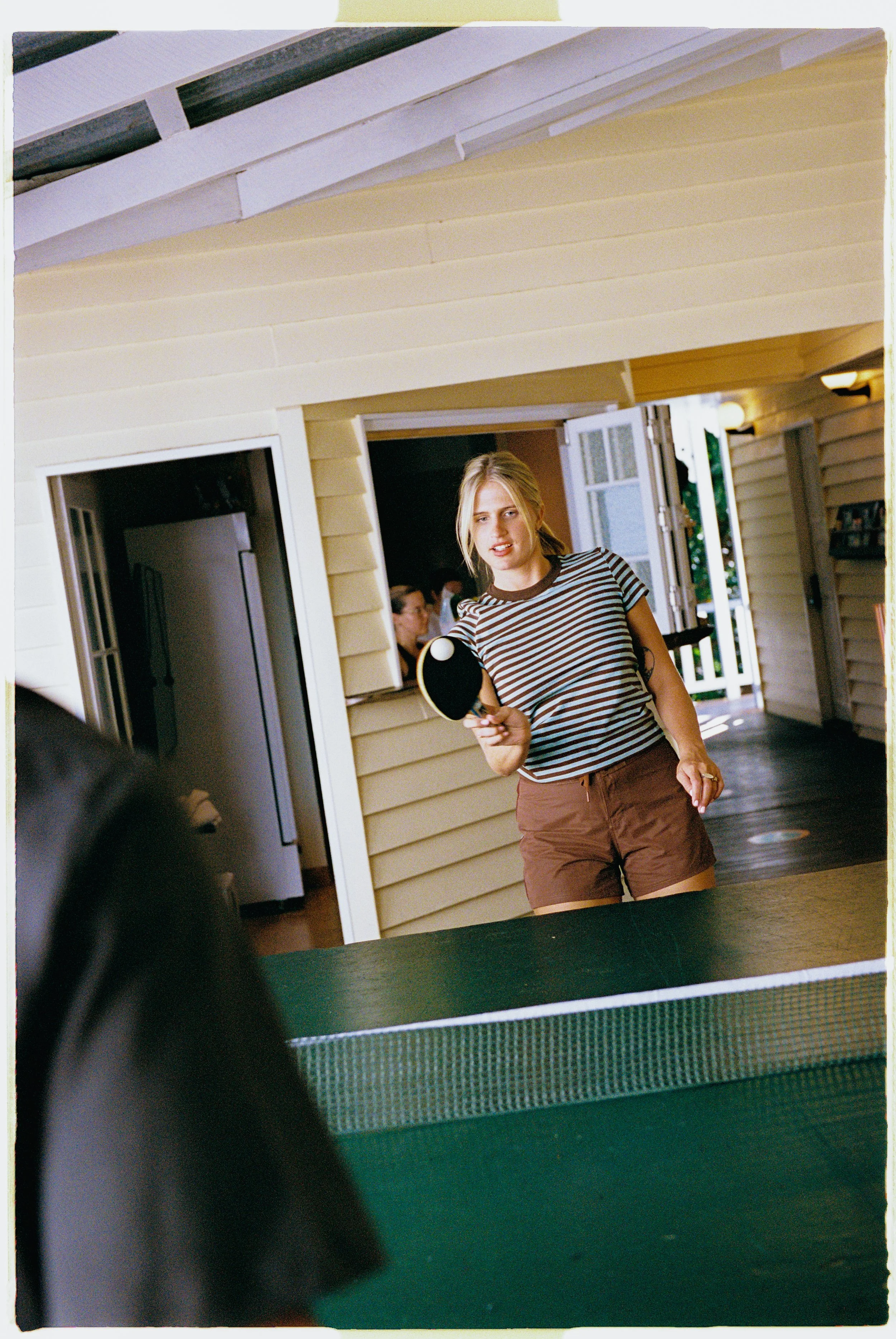 A young woman playing ping pong indoors, wearing a striped t-shirt and brown shorts, looking at the camera while holding a ping pong paddle.