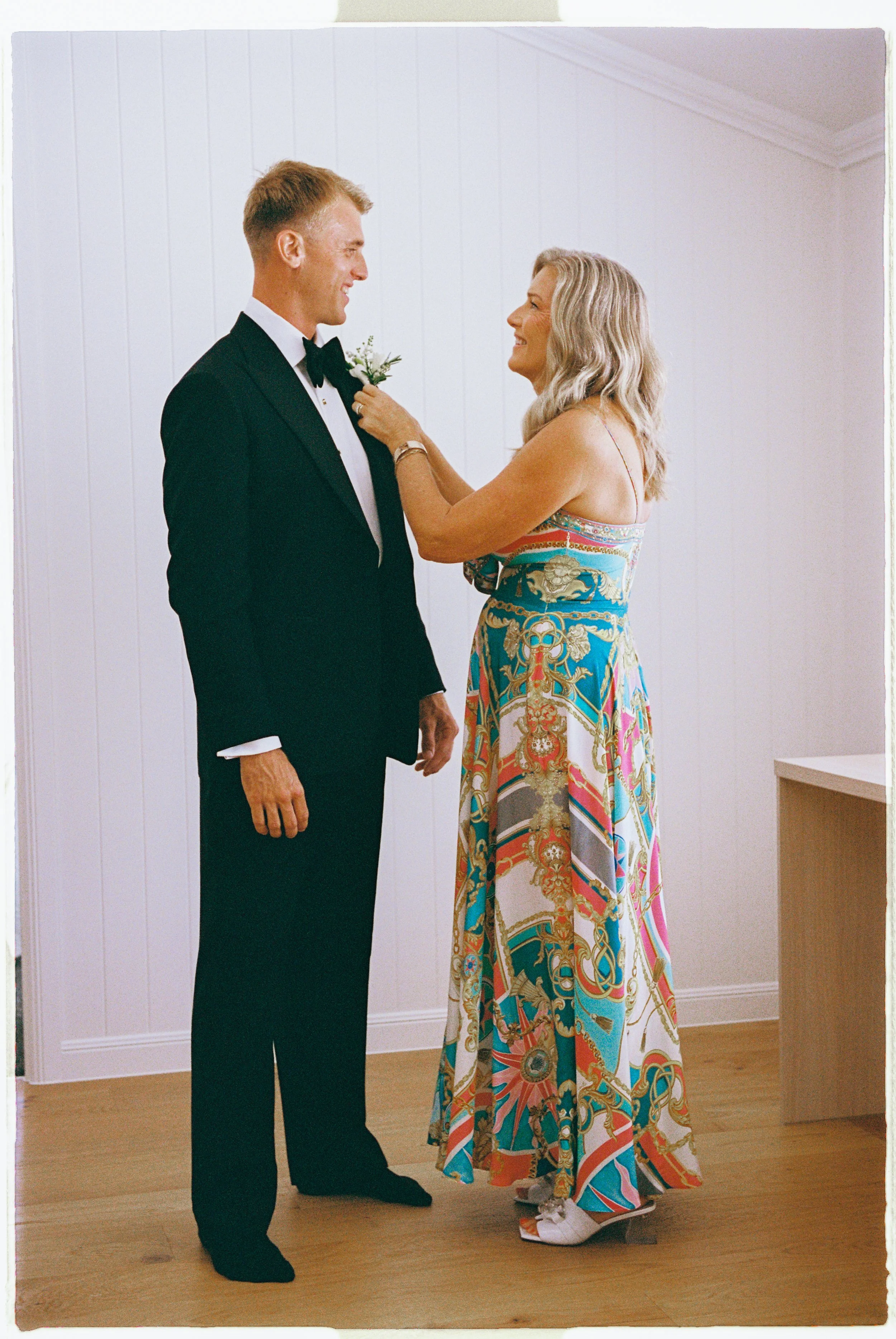A woman in a colorful, patterned dress pins a boutonniere on a man in a black tuxedo, both smiling, in a plain room with white walls.