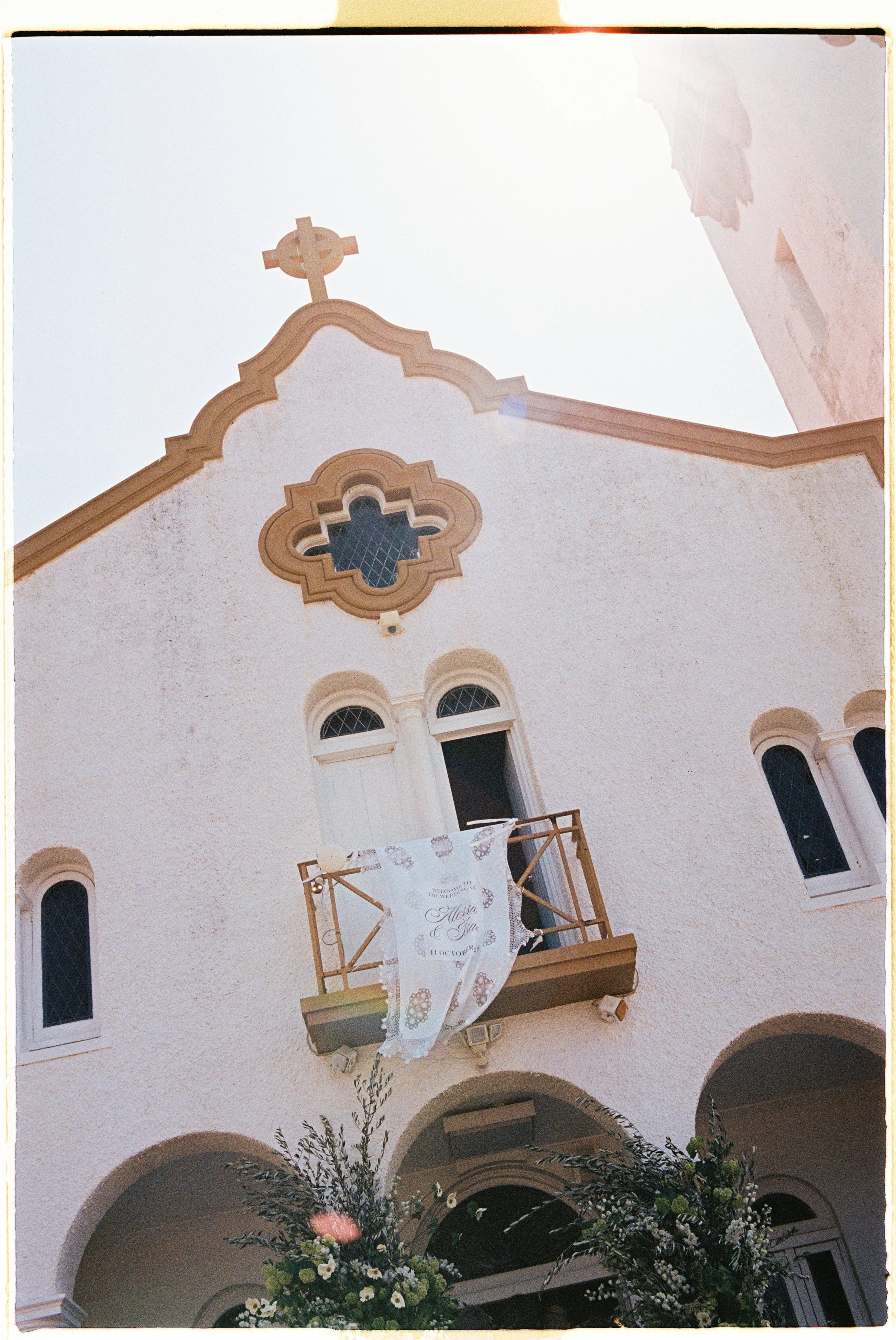 A small white church with arched windows and a cross on top. There is a balcony with a decorative cloth hanging and floral arrangements in the foreground.