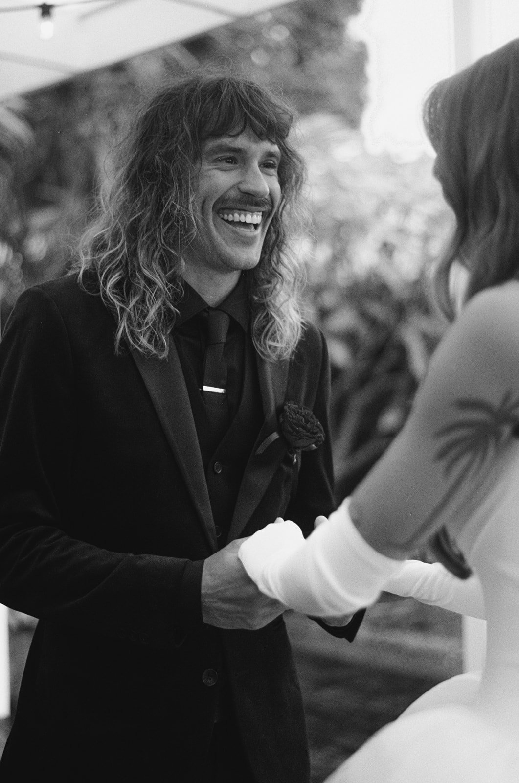 A person with long, curly hair, dressed in a dark suit and tie, smiling and shaking hands with a woman wearing a light-colored dress and gloves, at an outdoor event.