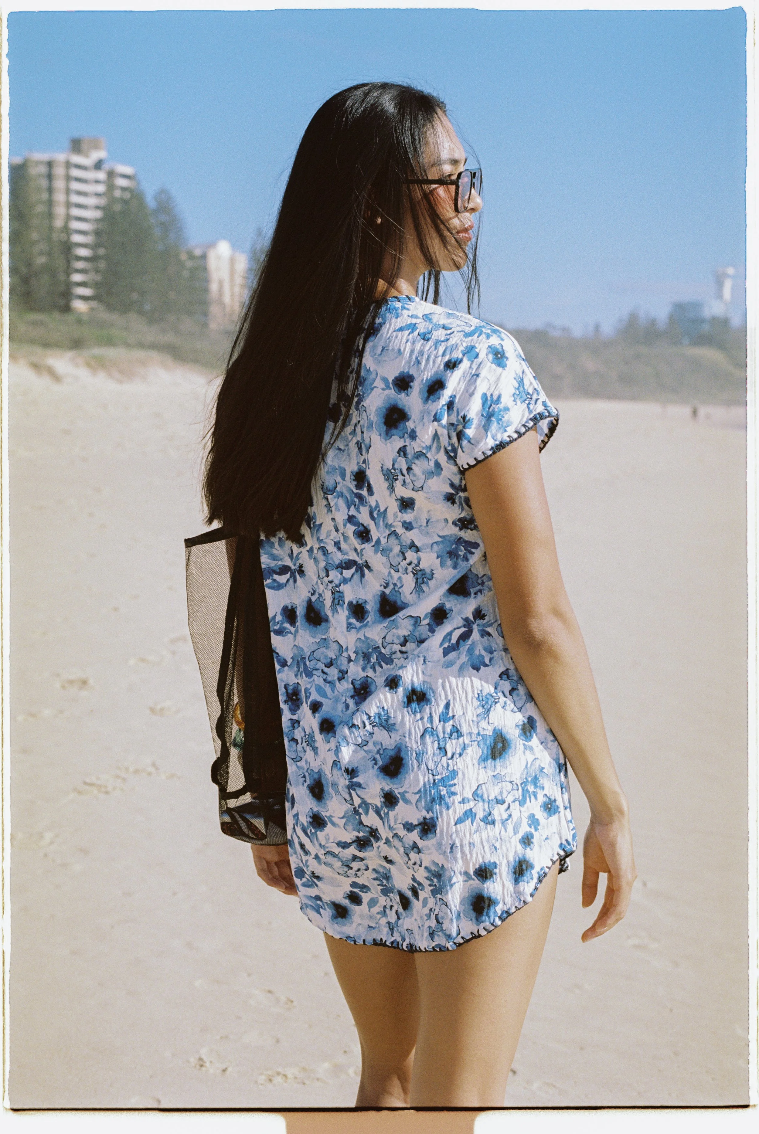 A woman in a blue floral dress and glasses standing on the beach, facing sideways with her hair blowing in the wind, with buildings and trees in the distance under a clear blue sky.