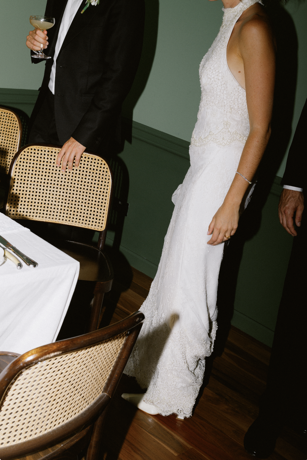 A bride and groom standing at a wedding reception table, with the groom holding a cocktail glass and the bride wearing a white lace wedding dress.