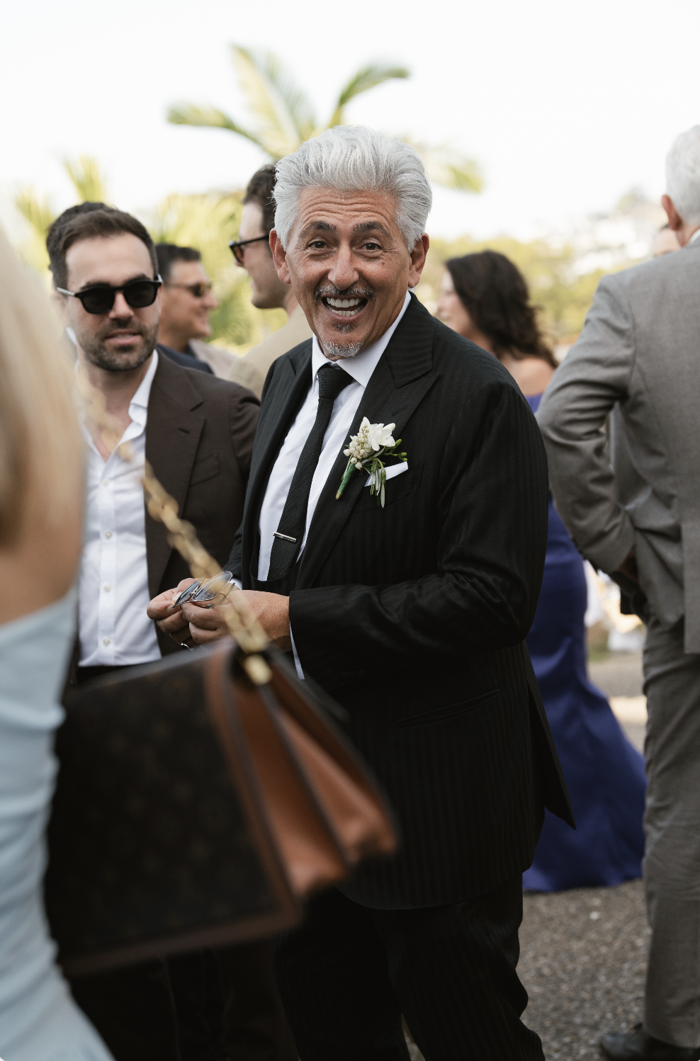 Smiling elderly man in a black suit and tie with a white boutonniere, party or wedding gathering outdoors.
