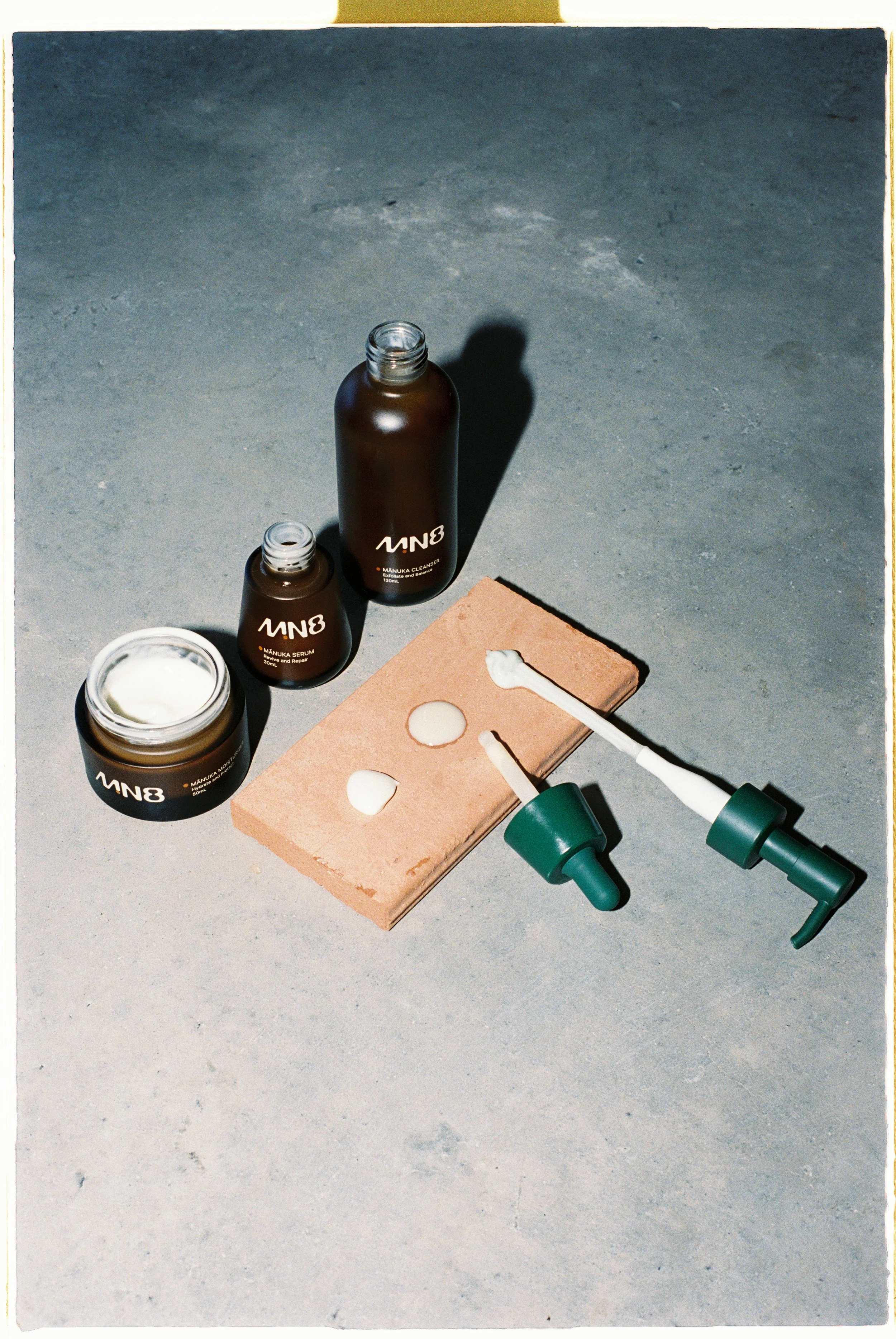 Skincare products including a jar, two bottles, and two droppers with green caps placed on a concrete surface with a brick in the background.