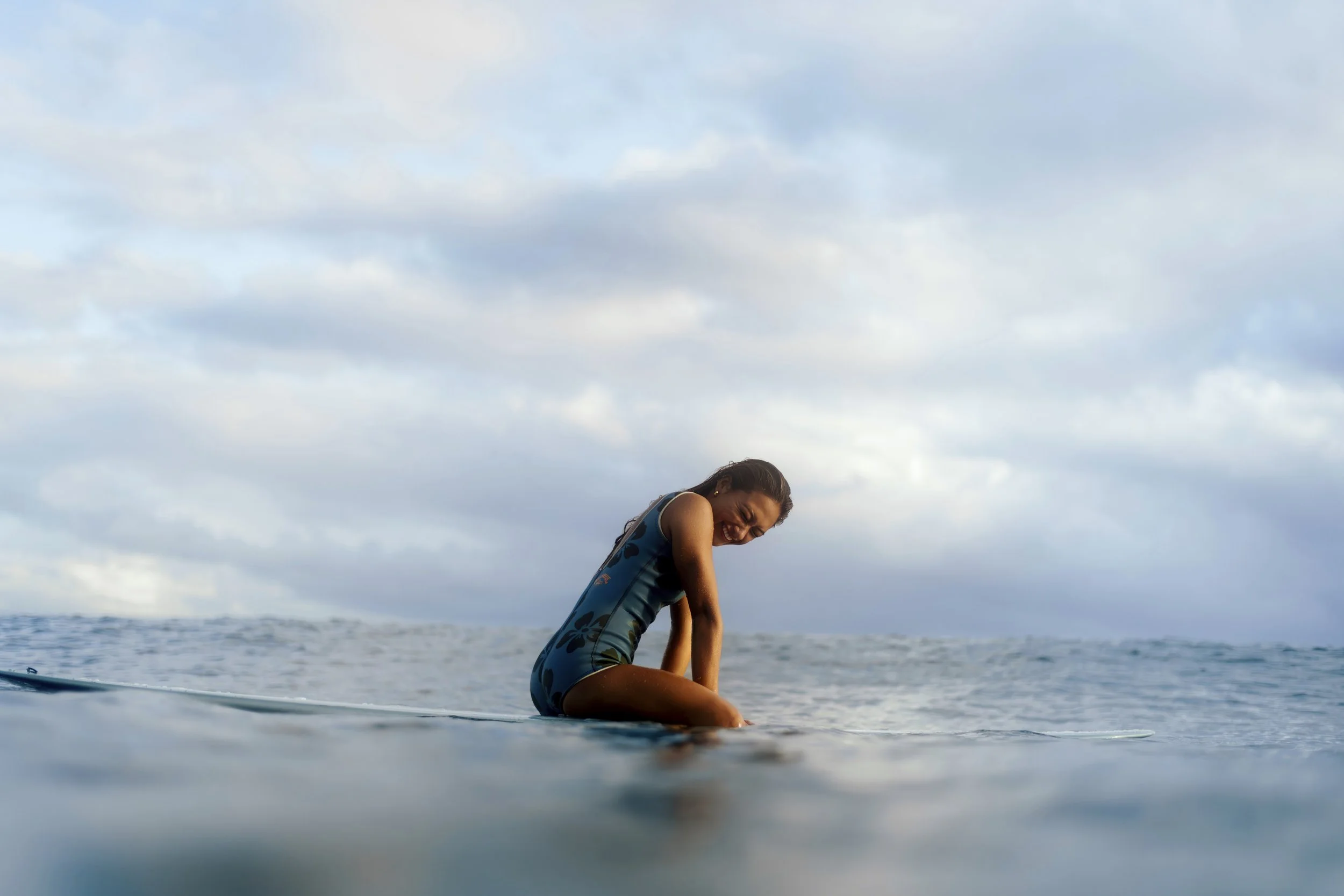 A woman sitting on a surfboard in the ocean, smiling and looking downward, under a cloudy sky.
