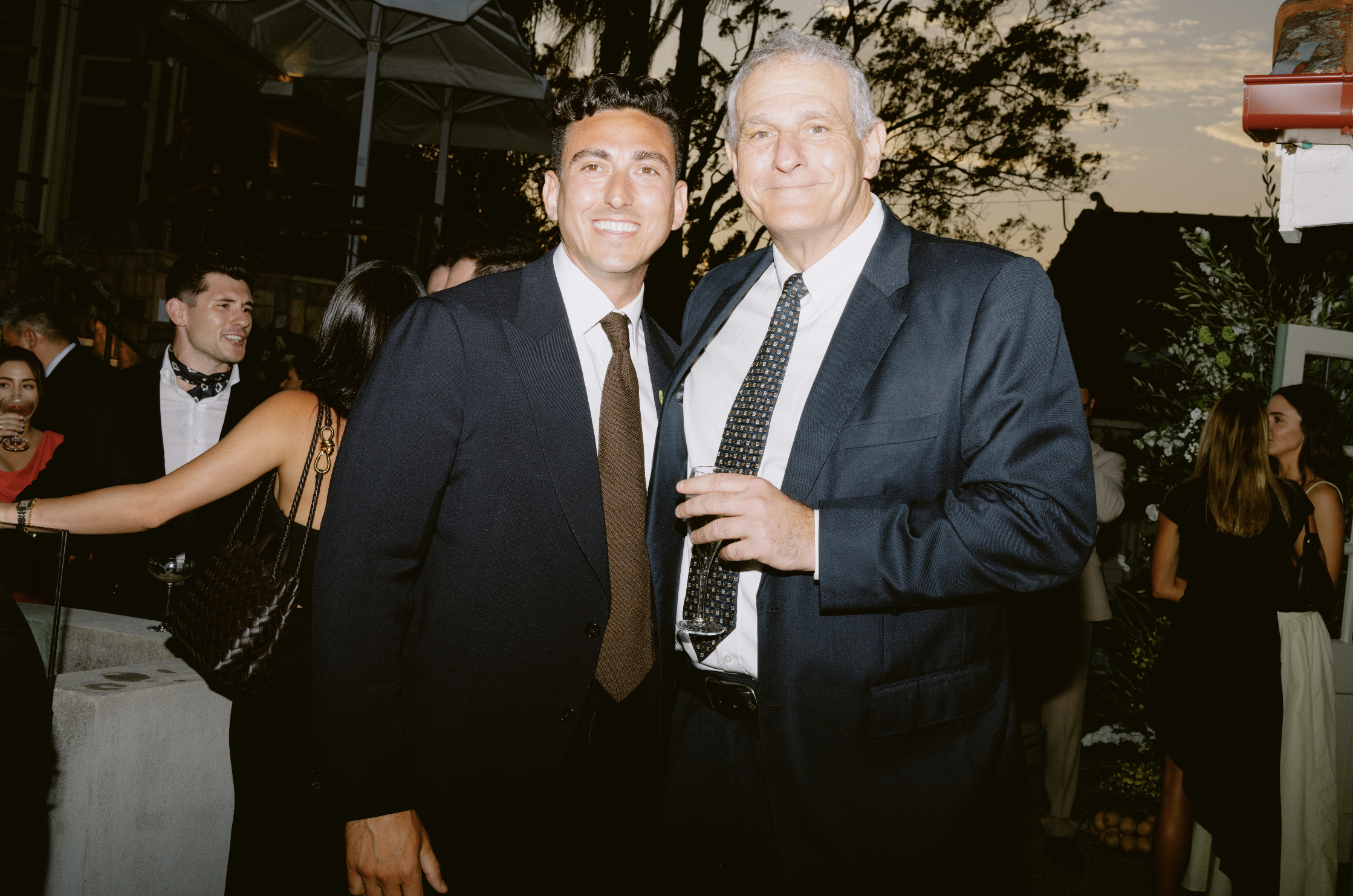 Two men in suits standing close together at an outdoor social event during sunset, with other guests mingling in the background.