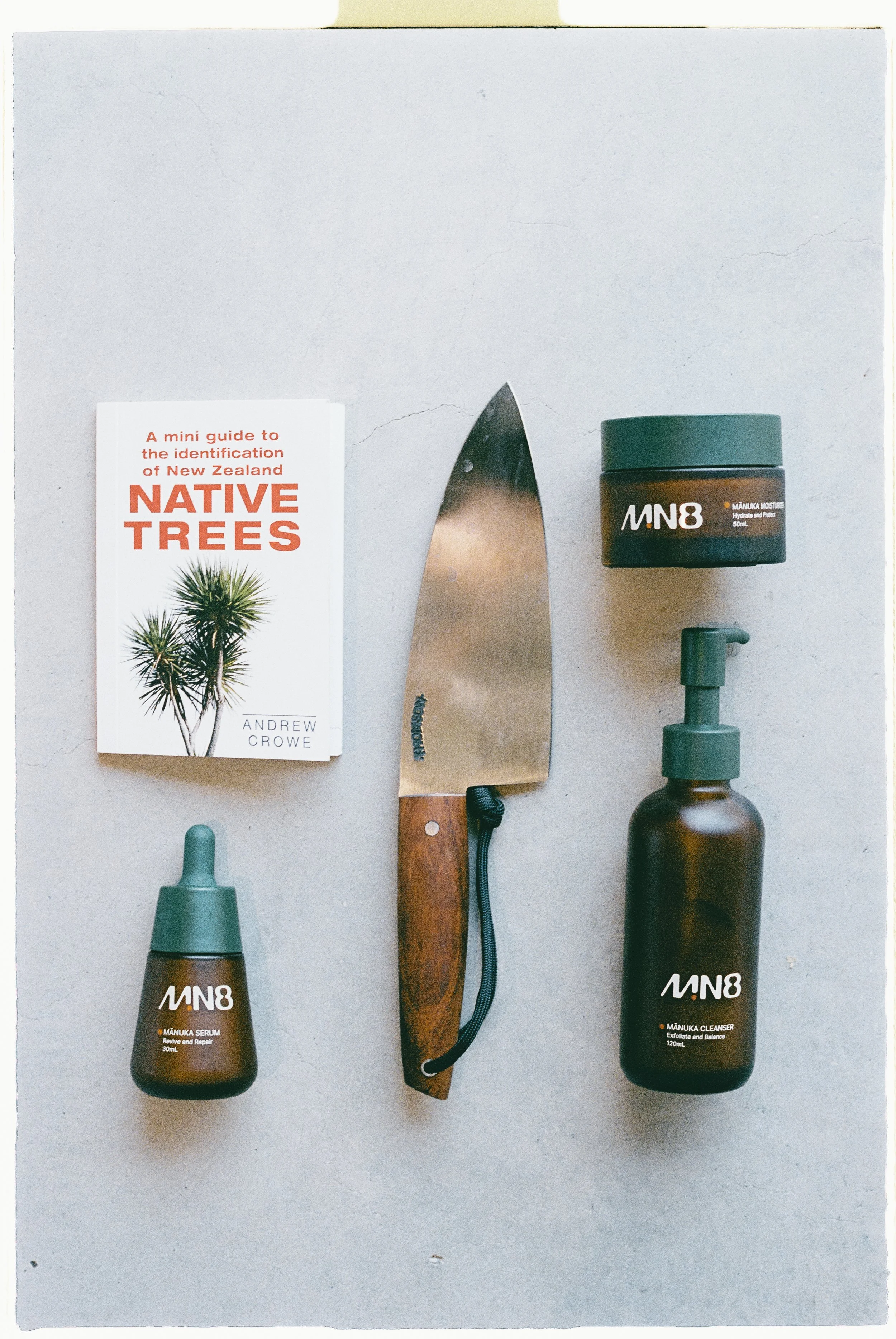 A flat lay of skincare products, a book titled 'A mini guide to the identification of New Zealand native trees' by Andrew Crowe, and a pocket knife with a wooden handle on a light gray surface.