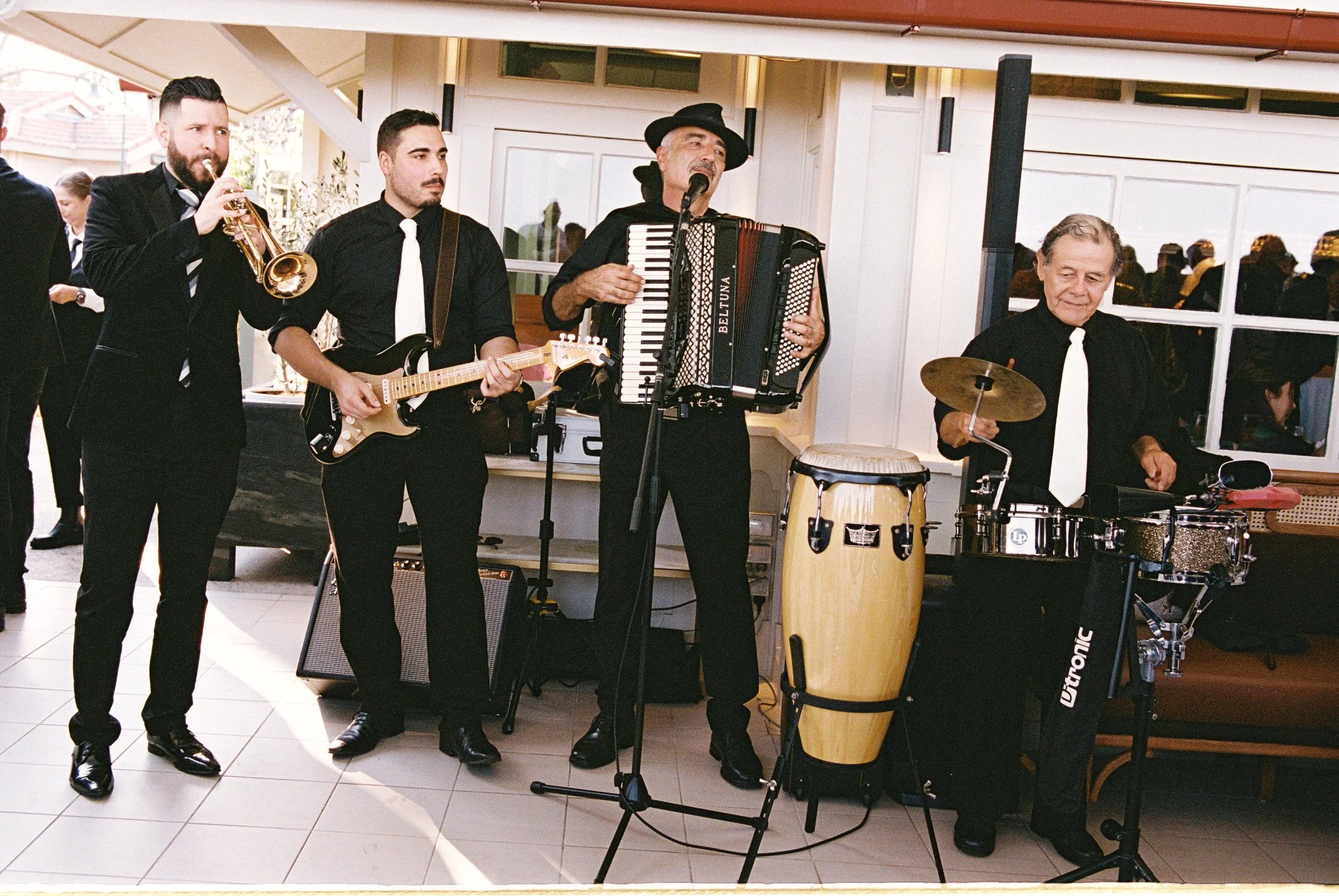 A band performing music at an indoor event, featuring five musicians playing a trumpet, electric guitar, accordion, conga, and drum.