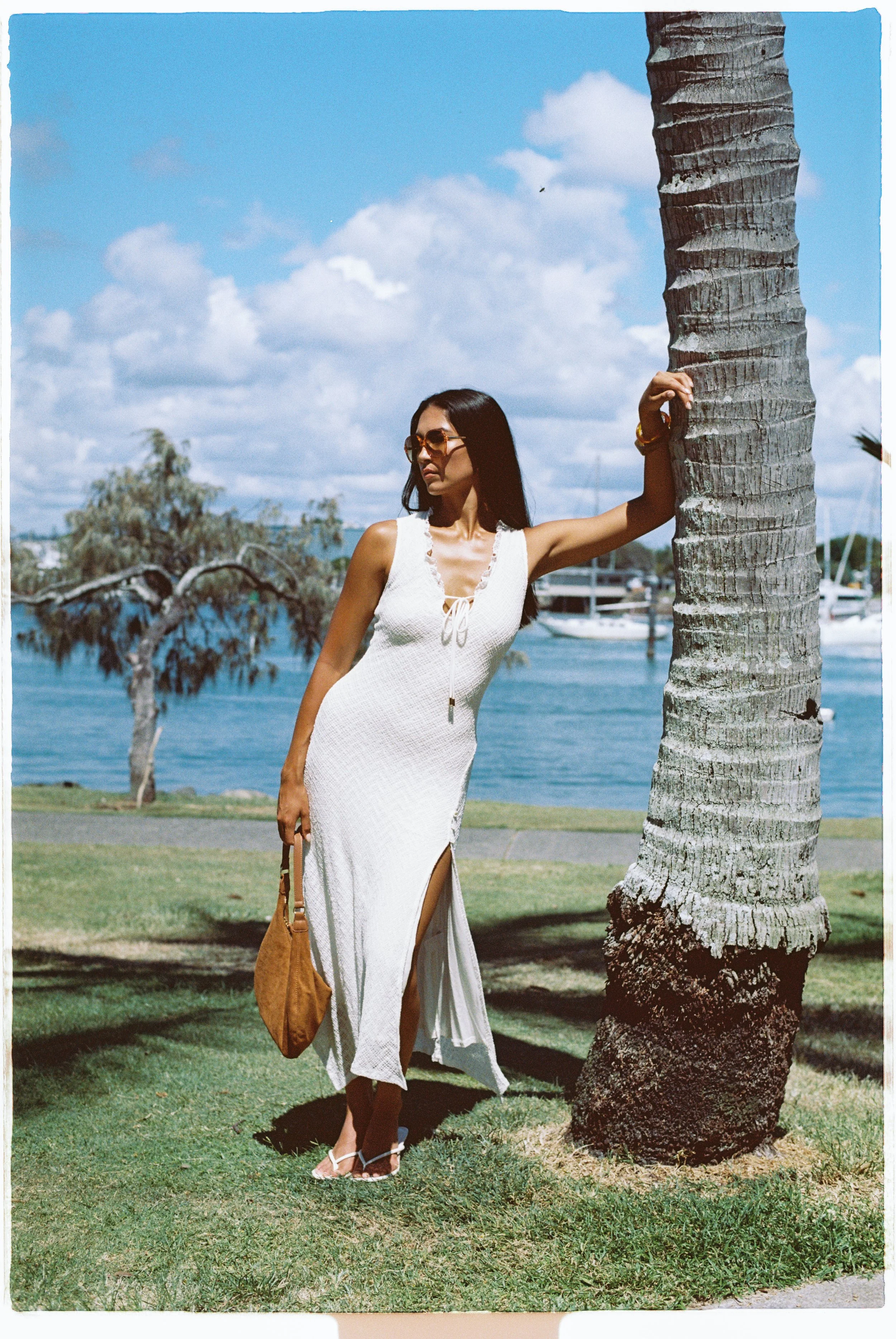 A woman in a white sleeveless dress with a slit, sunglasses, and beige handbag standing under a palm tree near the water with boats and sailboats in the background.