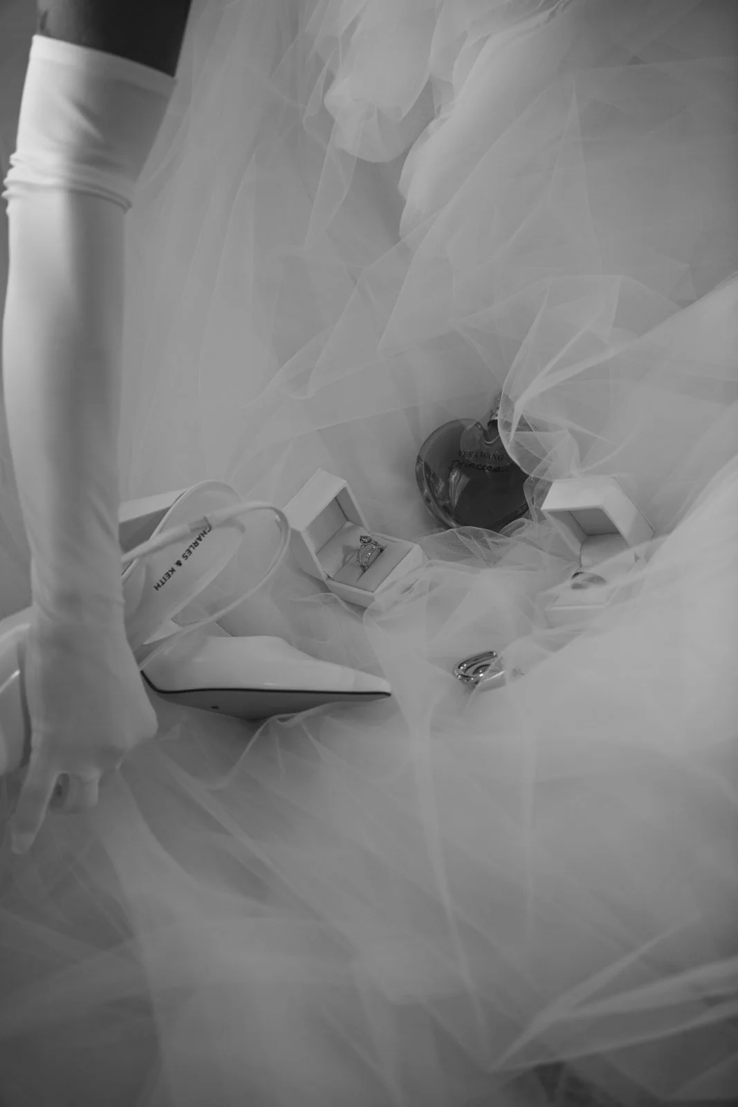 A white high-heeled shoe, rings in a box, a perfume bottle, a ring, and a bracelet displayed on white fabric, likely for a wedding or special occasion.