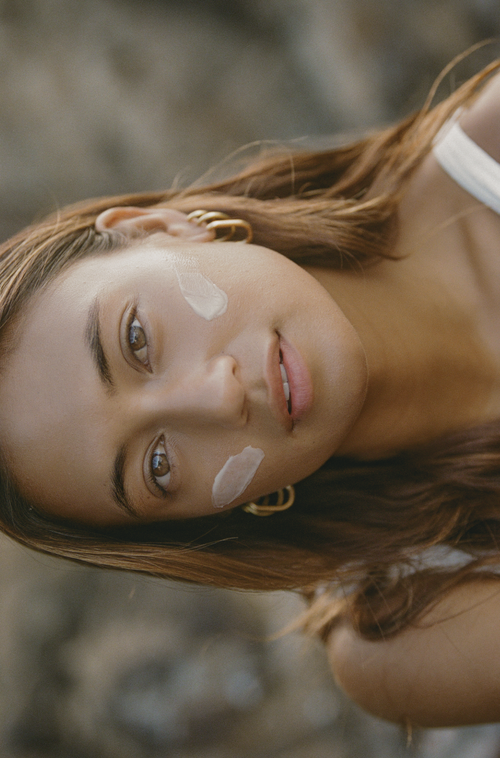Close-up of a young woman with light brown hair, lying down on a rocky surface, with cream on her face and gold hoop earrings.