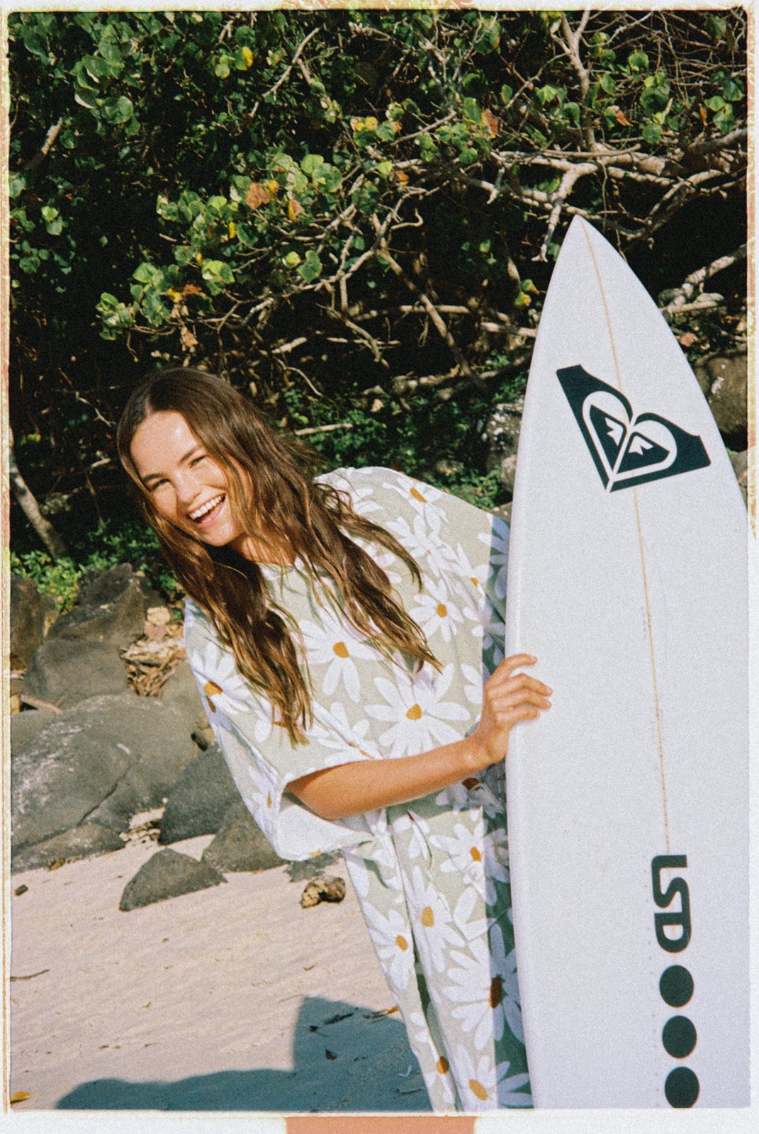 A young woman with long brown hair, wearing a daisy print jumpsuit, holding a surfboard, and smiling at the camera on a sandy beach with rocks and greenery in the background.