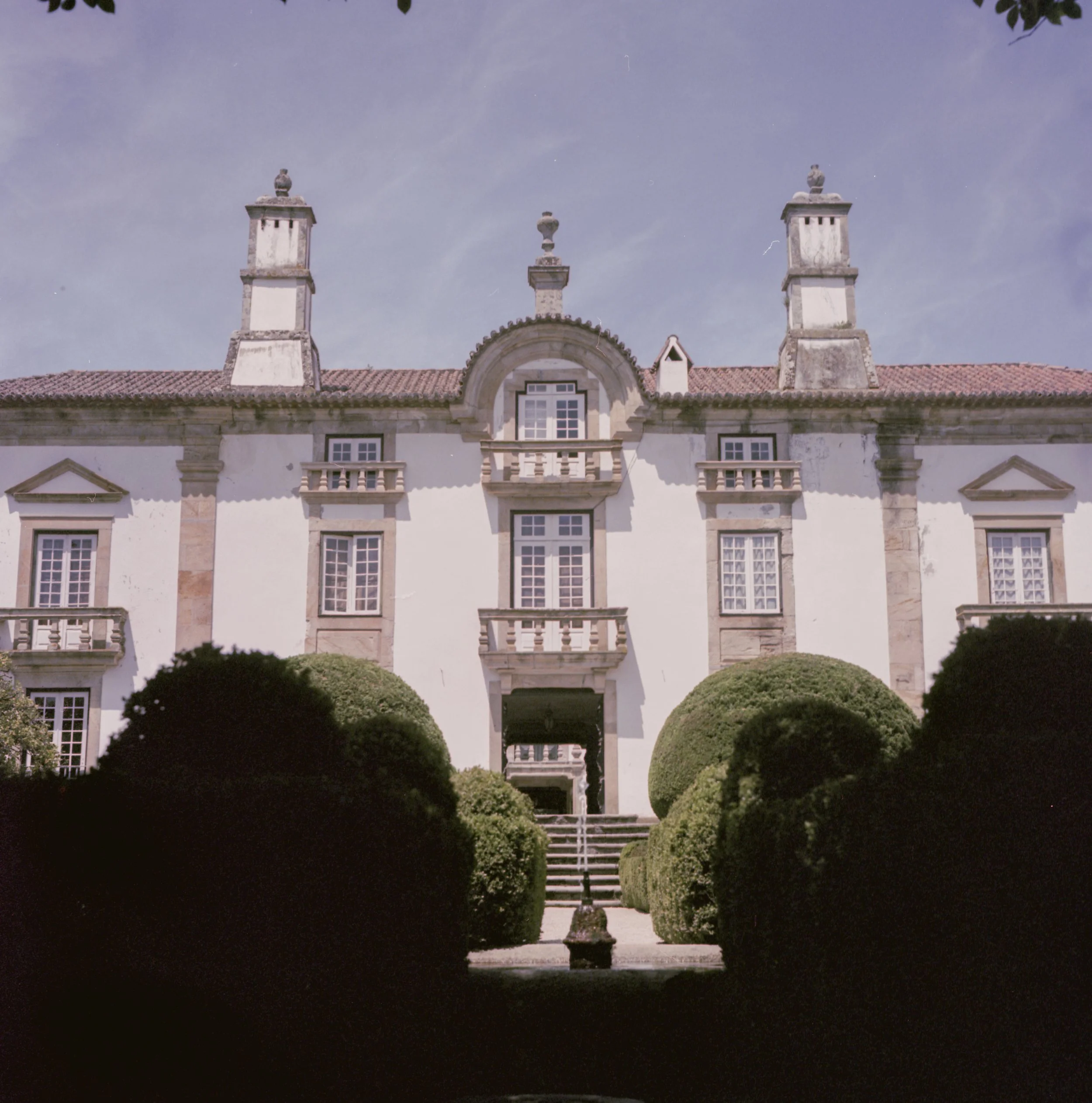 A grand historic building with a symmetrical facade, multiple windows, and a roof with decorative chimneys or spires. The building has an arched entrance and well-maintained hedges in the front yard. The sky above is clear with some faint wisps of cl