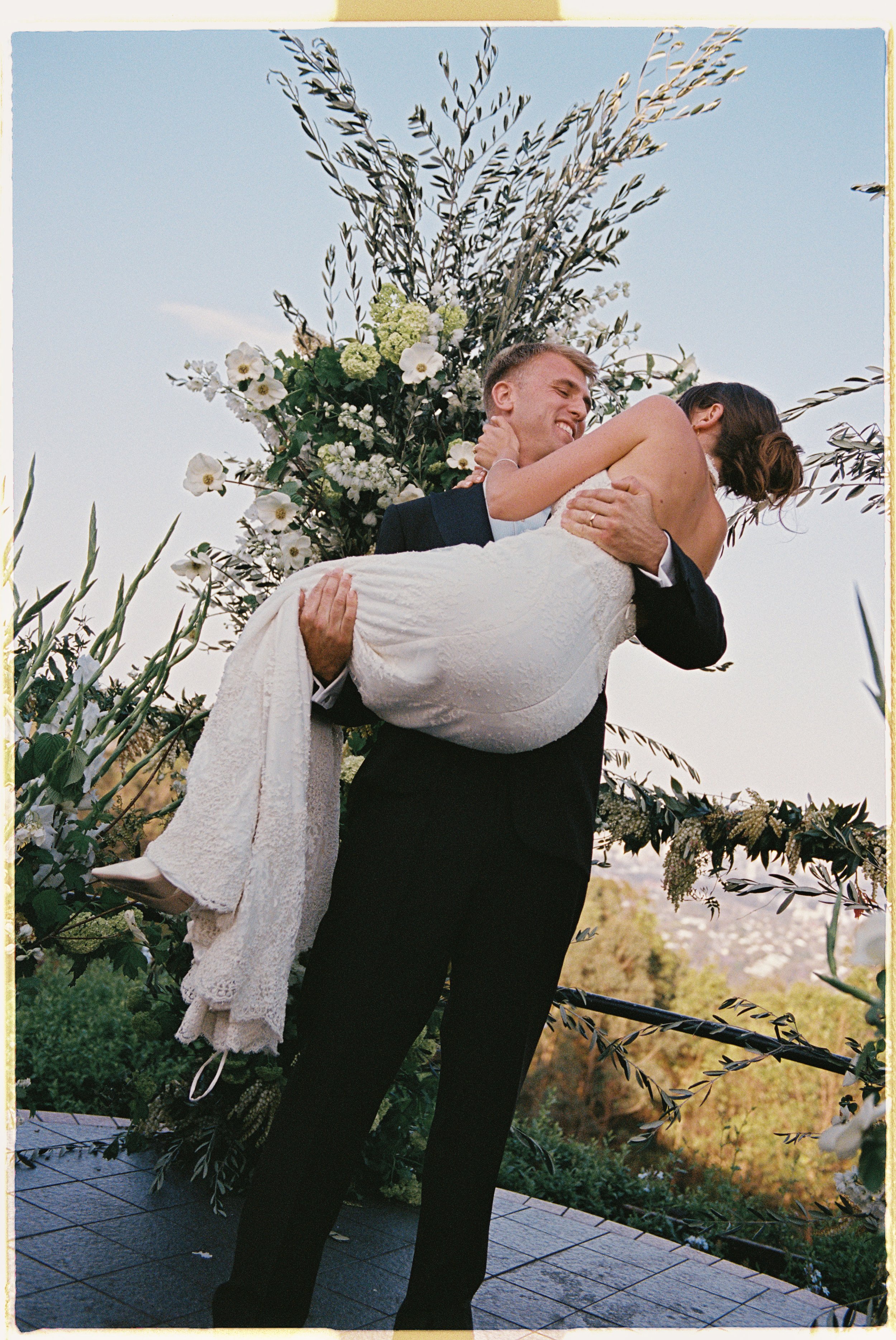 A groom in a black suit holding a bride in a white lace wedding dress, lifting her in the air with a large floral arrangement in the background outdoors.