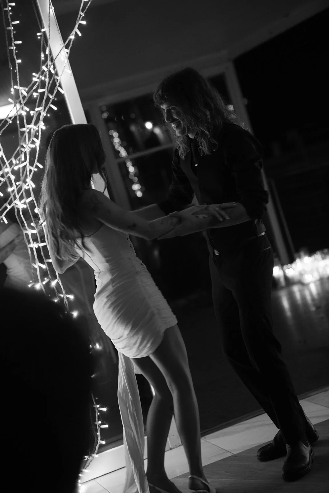 A woman in a white dress appears to be receiving a gift from another woman in dark clothing, with string lights in the background.