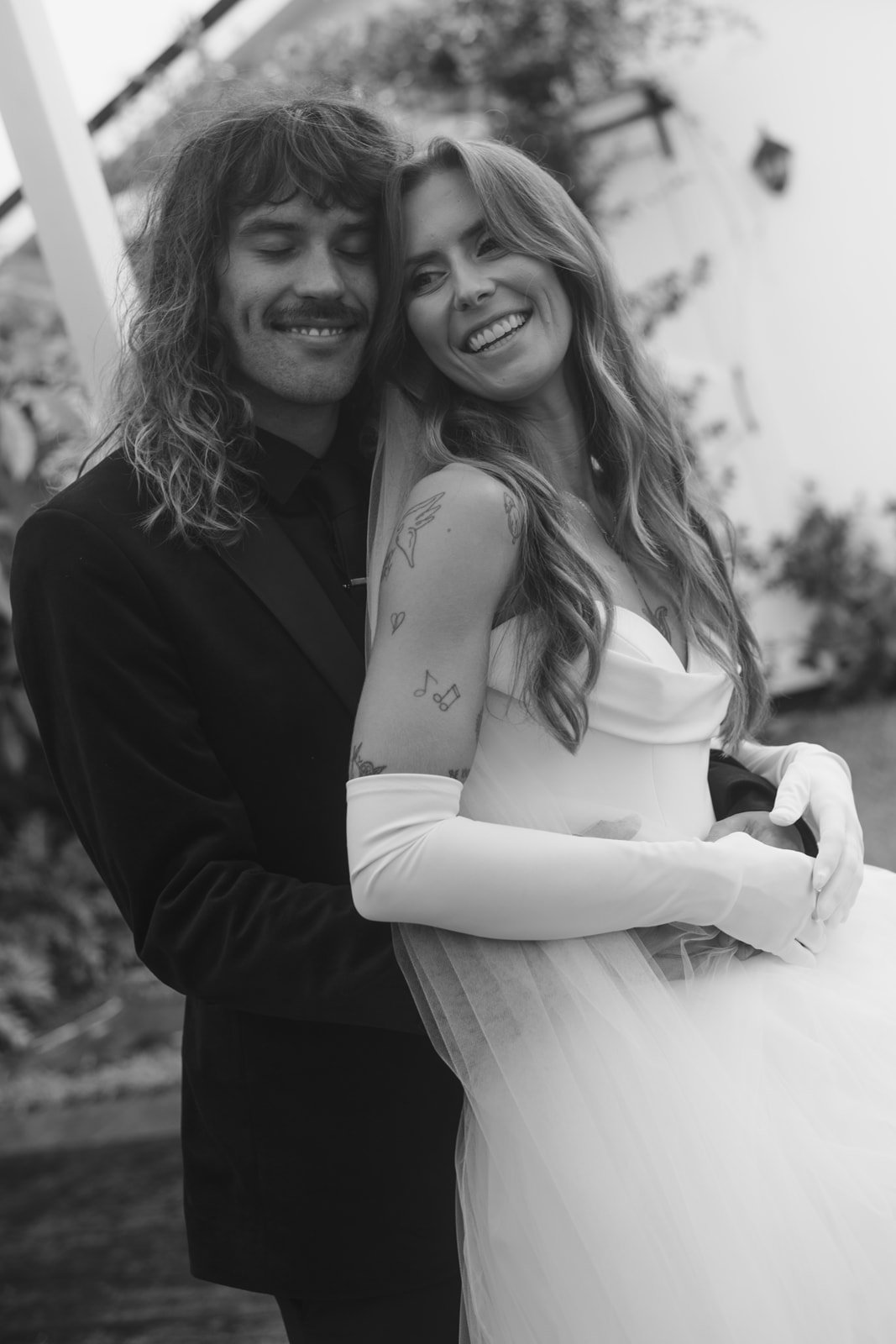 A smiling couple on their wedding day, with the man in a dark suit and the woman in a strapless wedding gown and satin gloves, posing outdoors.