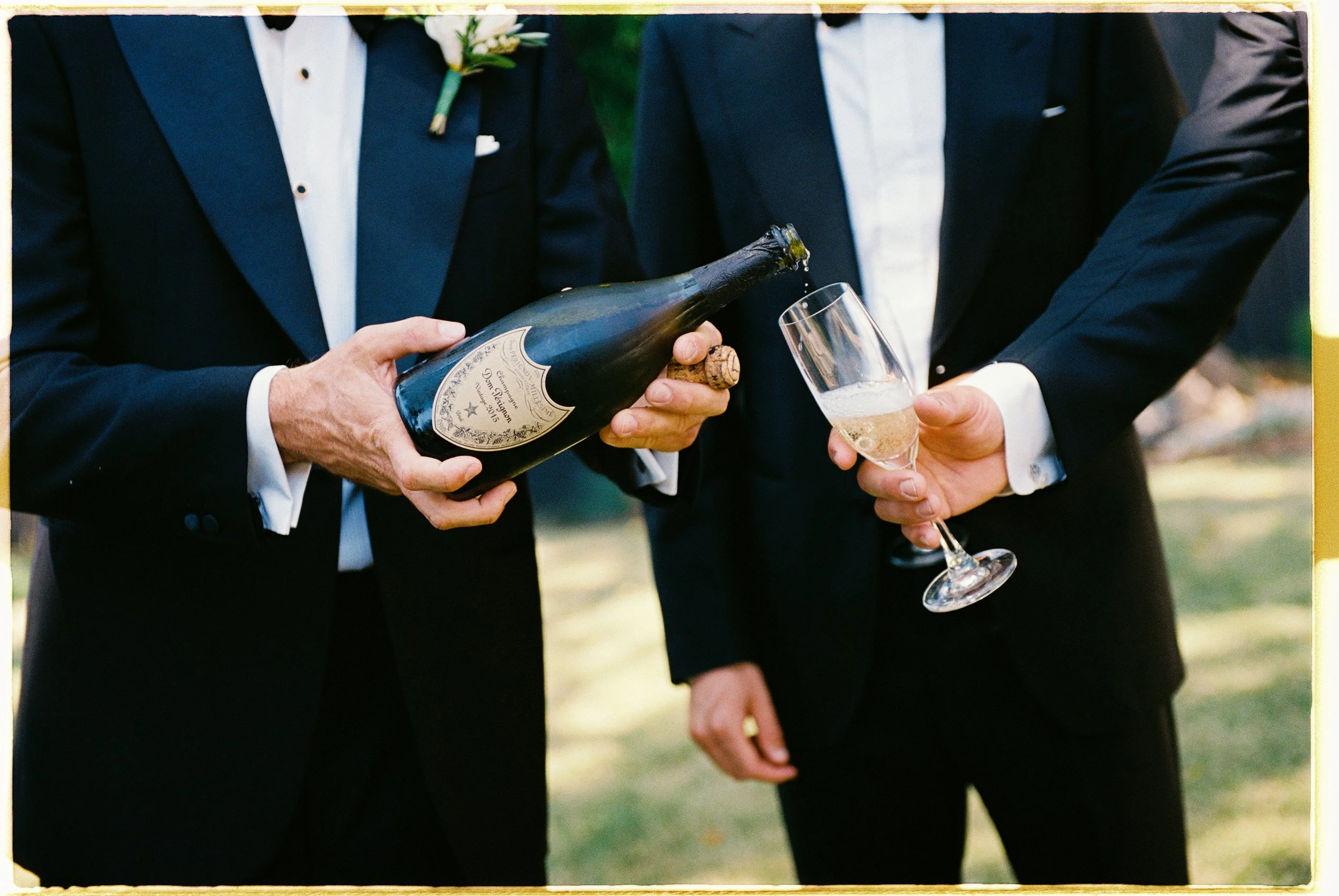 Two men in tuxedos hold a bottle of champagne and a champagne flute at an outdoor event.