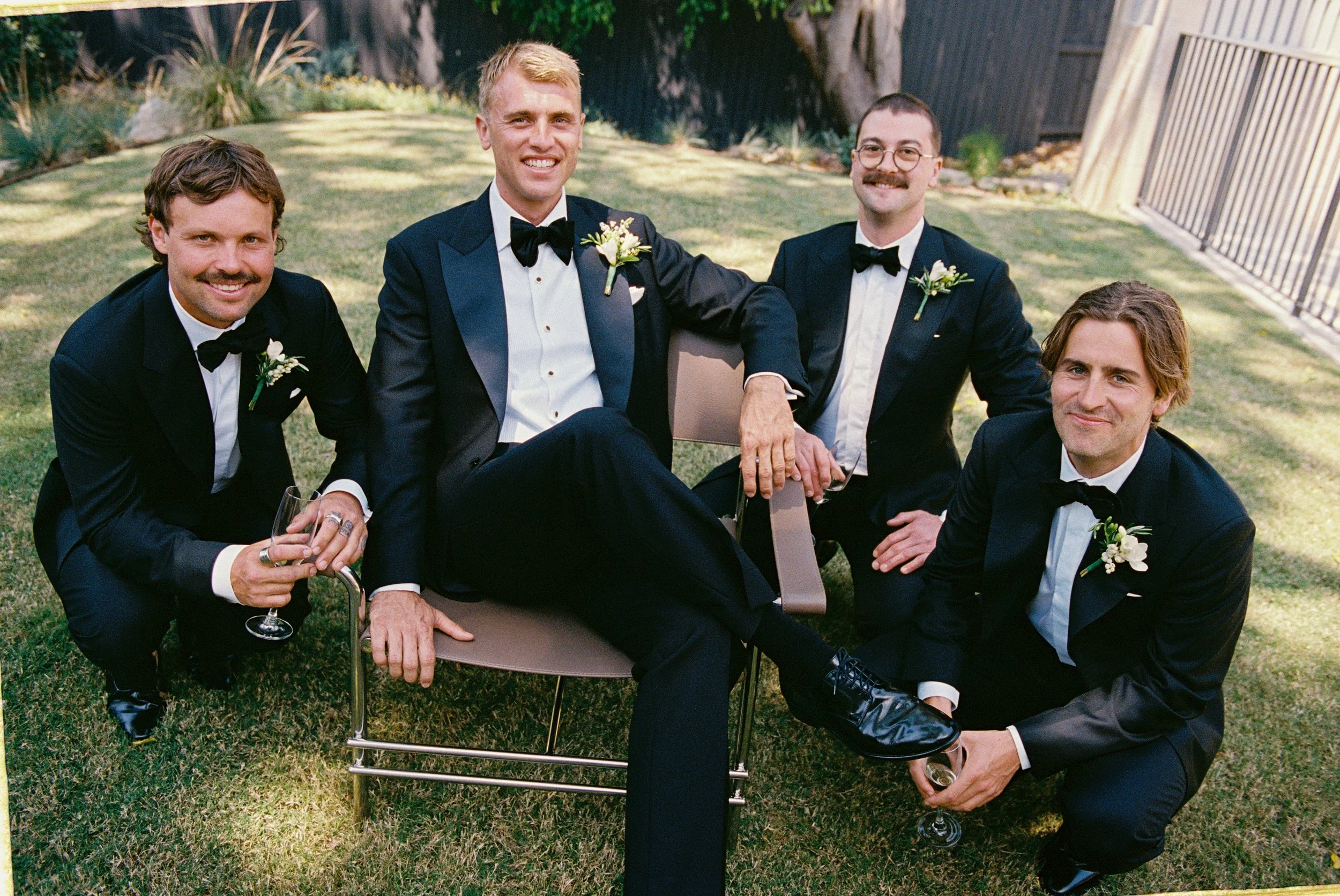 Five men dressed in black tuxedos with bow ties and boutonnières, outdoors on a grassy lawn, sitting and kneeling, holding glasses of champagne