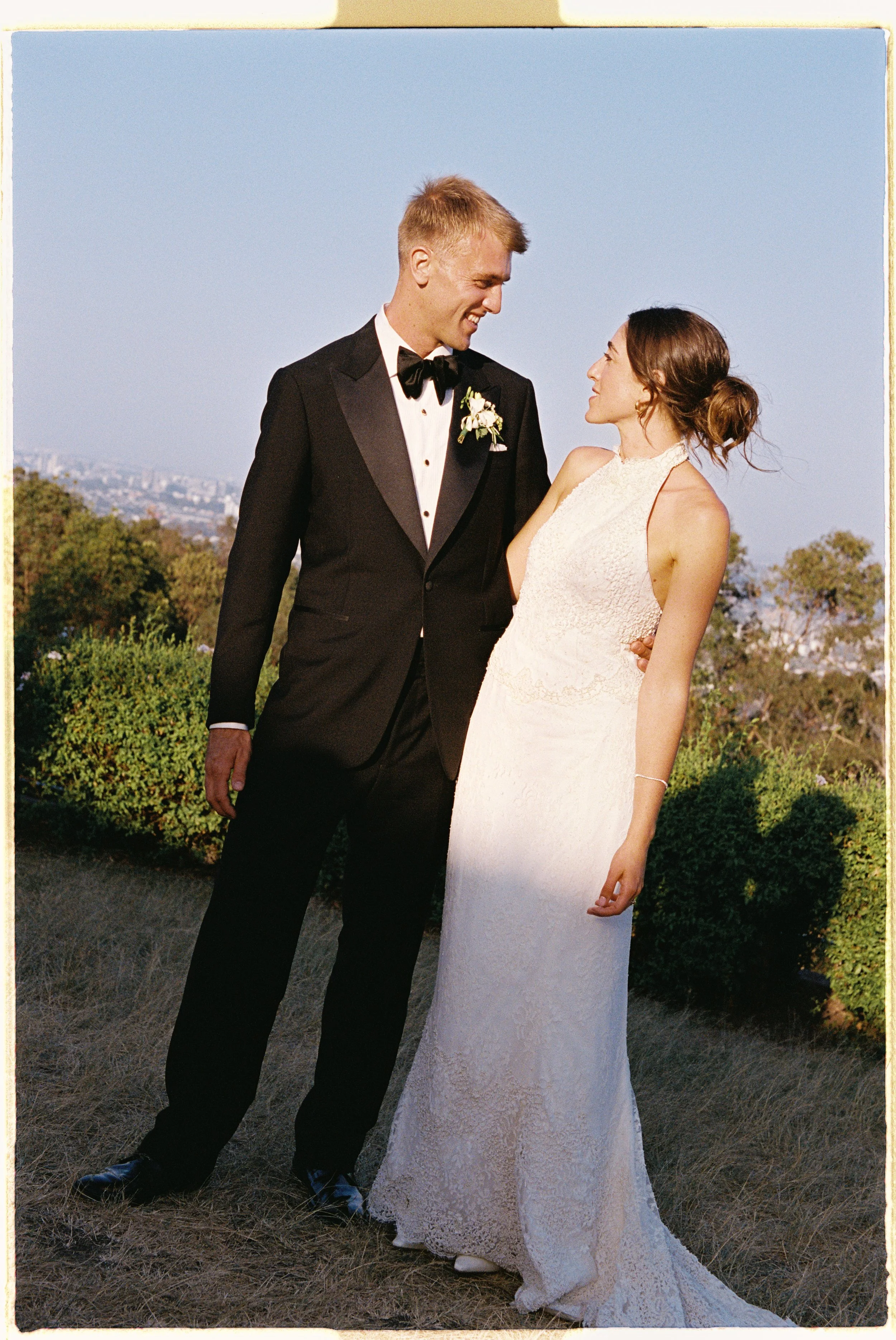 A bride and groom dressed in wedding attire standing outdoors on a grassy area with trees and a city skyline in the background, smiling at each other during sunset.