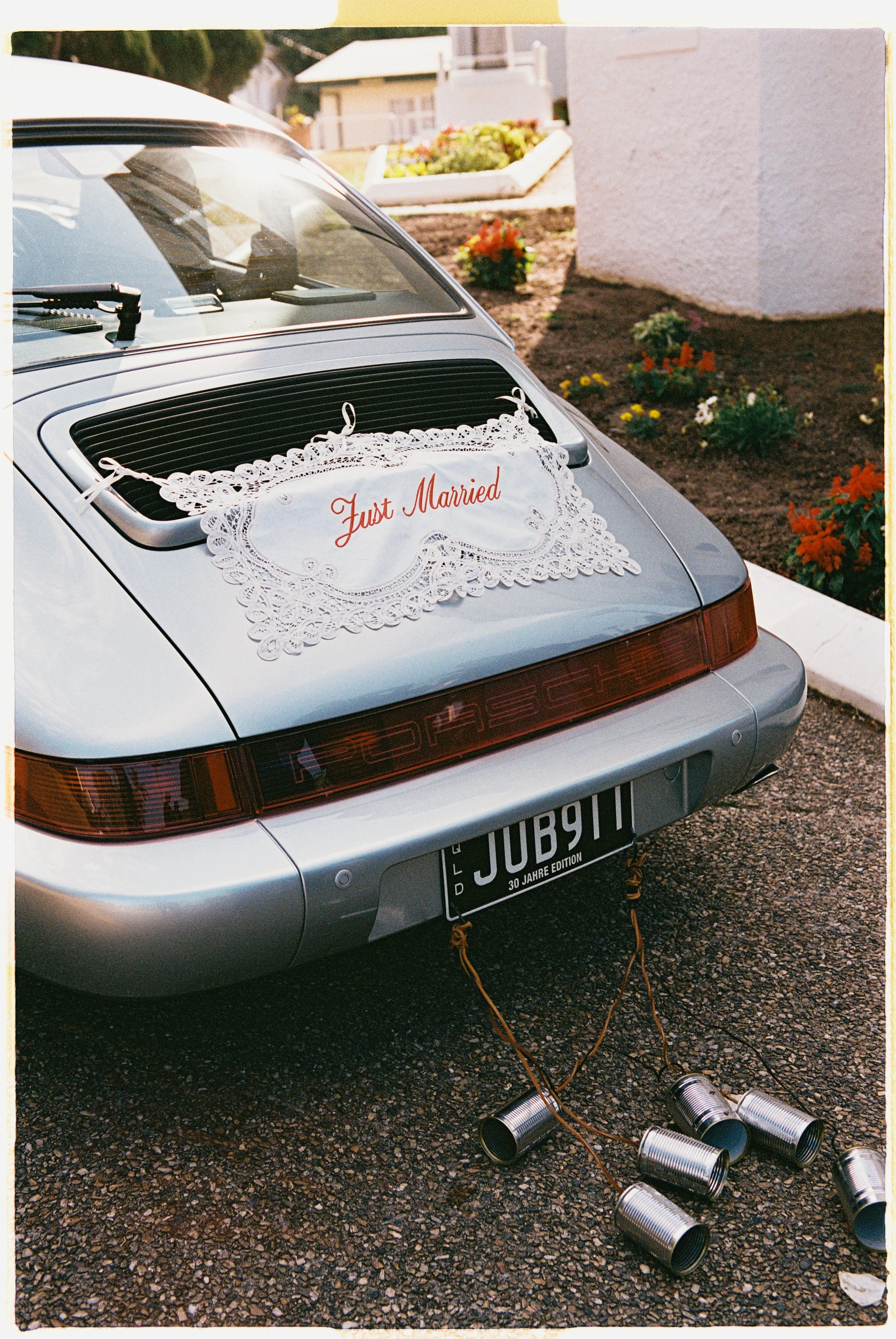 The rear of a silver Porsche 911 with a lace doily on the back window that has red embroidered text reading 'Just Married.' The car is decorated for a wedding and is parked on a gravel driveway with empty tin cans tied to the bumper by strings. There