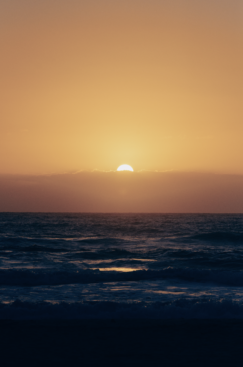 Sunset over the ocean with the sun partially hidden behind clouds and calm waves.