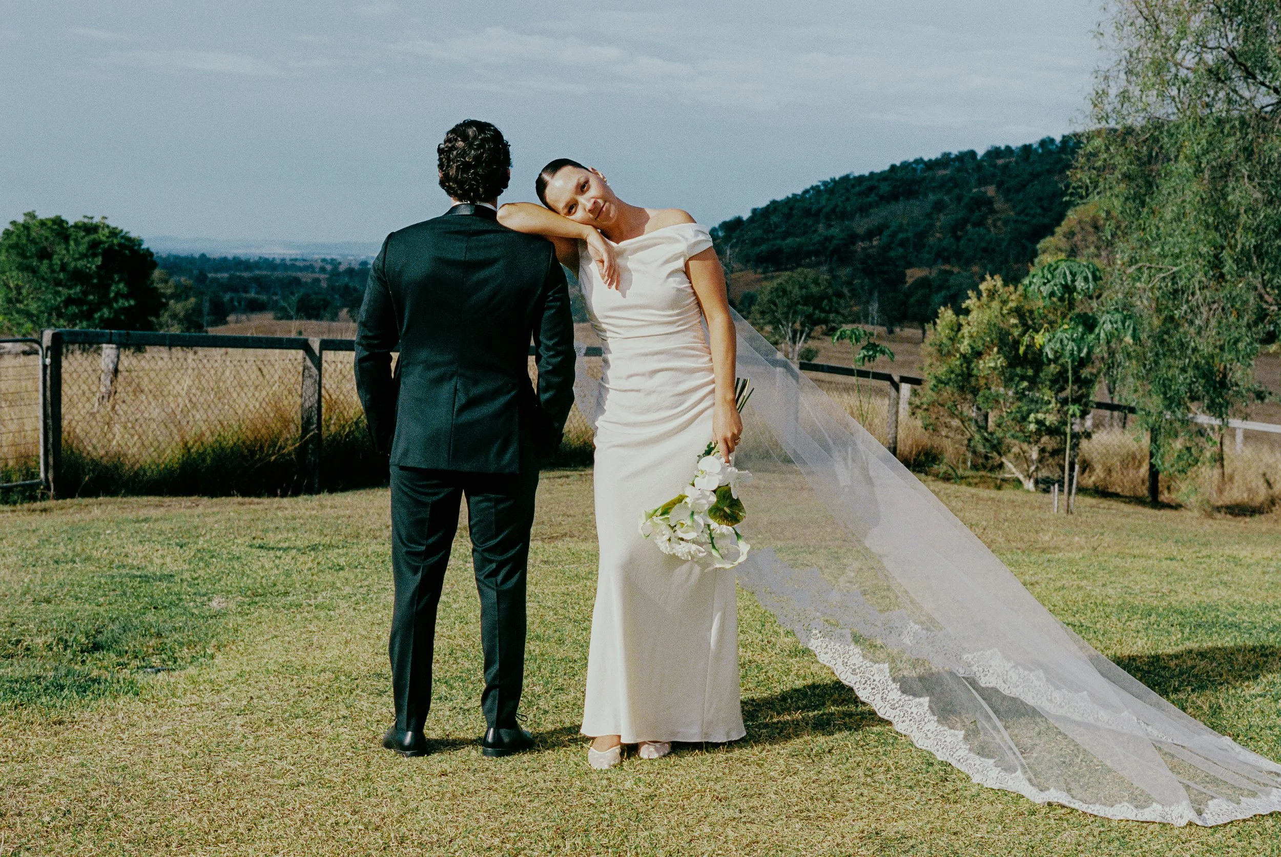A bride in a white wedding dress with a lace train, holding a bouquet of white flowers, leans her head on a groom dressed in a black suit, standing outdoors on a grassy area with trees, a fence, and hills in the background.