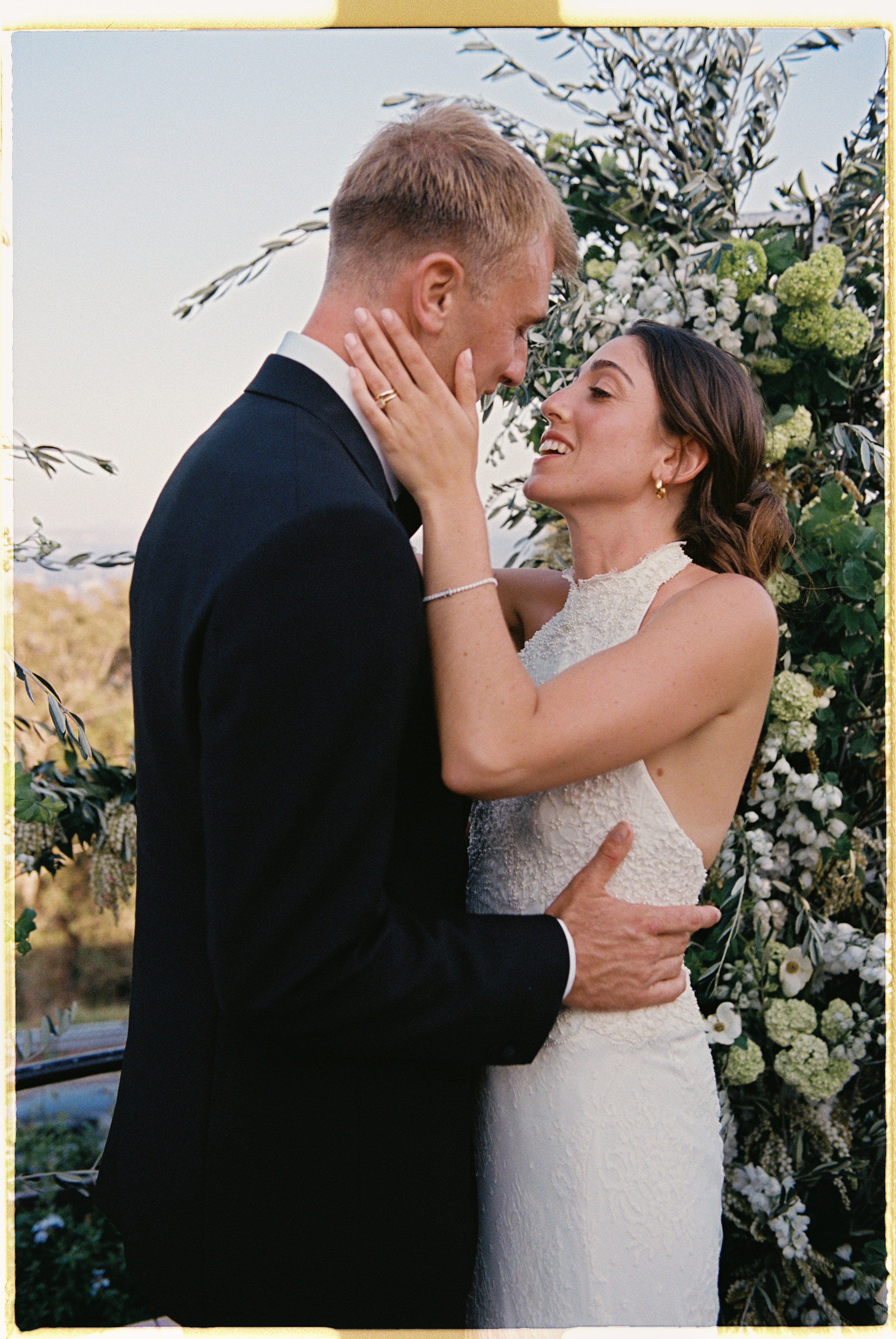 A bride and groom embrace outdoors during their wedding, with the bride holding the groom's face and the groom holding her waist, surrounded by bridal floral arrangements.