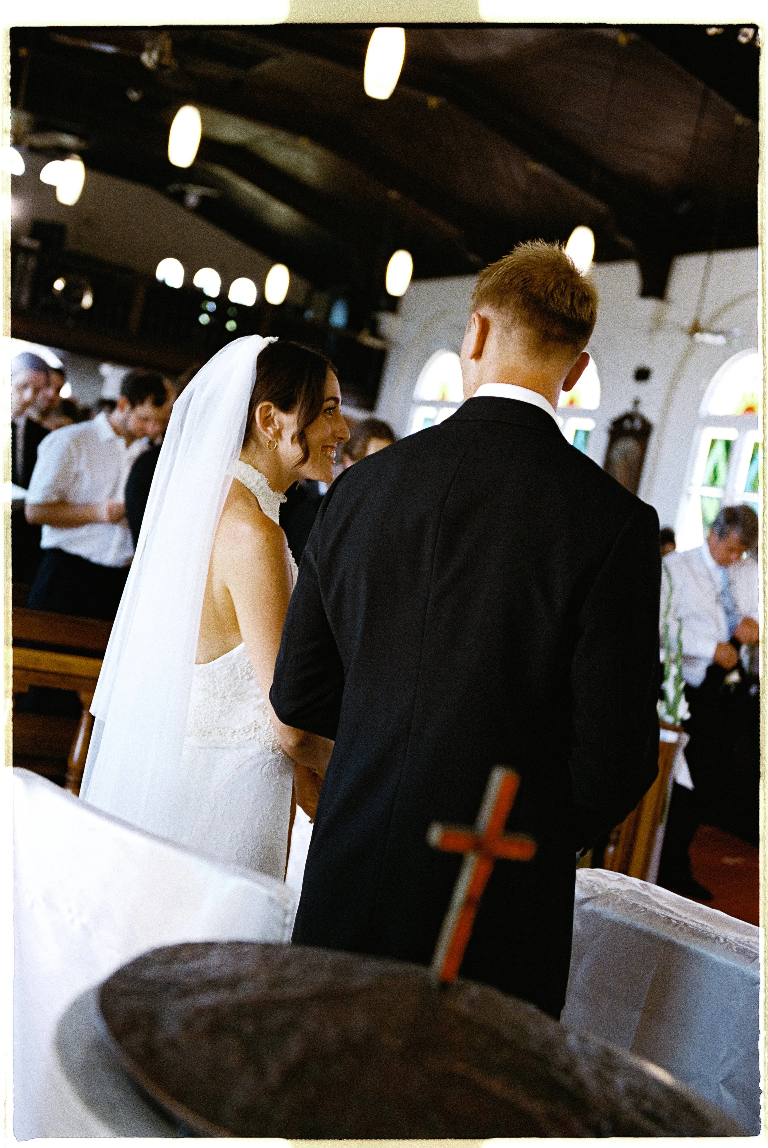Bride and groom standing in church during wedding ceremony, with guests in the background and wooden ceiling with hanging lights.