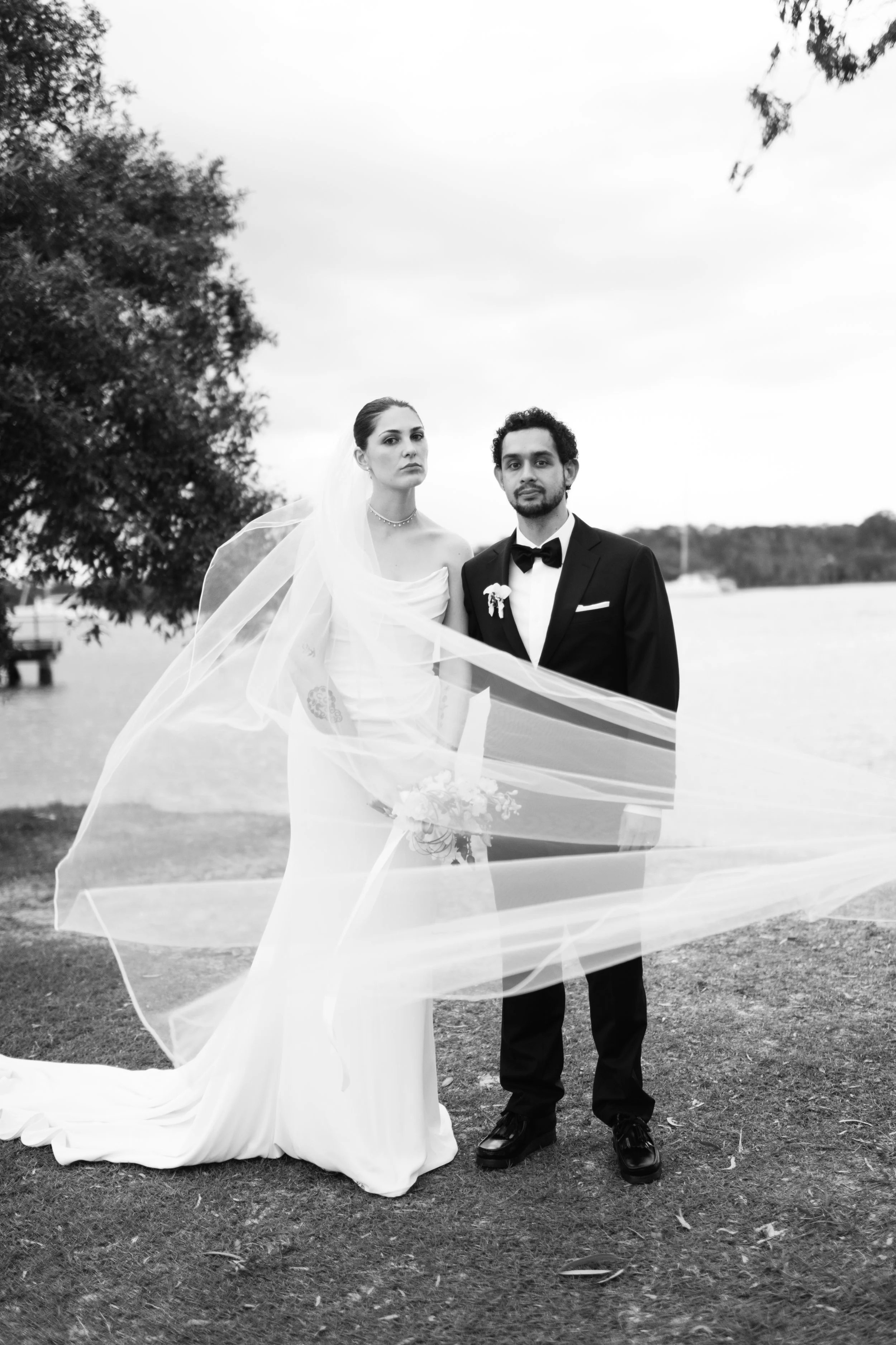 A black and white photo of a bride and groom outdoors. The bride wears a wedding dress with a veil, and the groom wears a tuxedo with a bow tie. They stand on grass with trees and a body of water in the background.