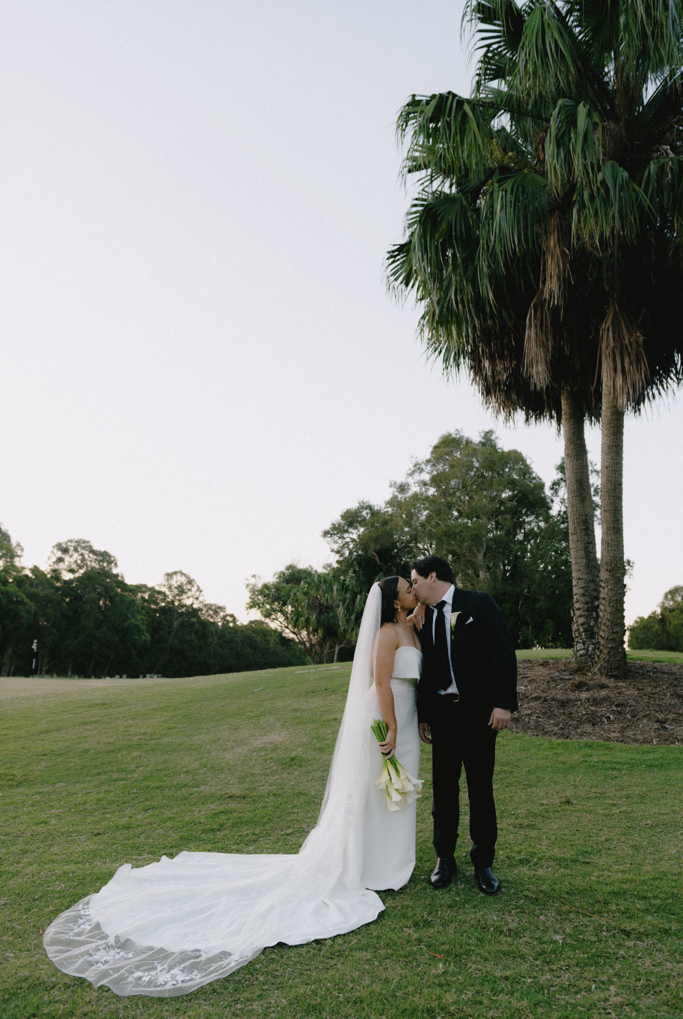 Bride and groom kissing outdoors on a grassy field next to palm trees, the bride holding a bouquet of white calla lilies, at sunset or early evening.