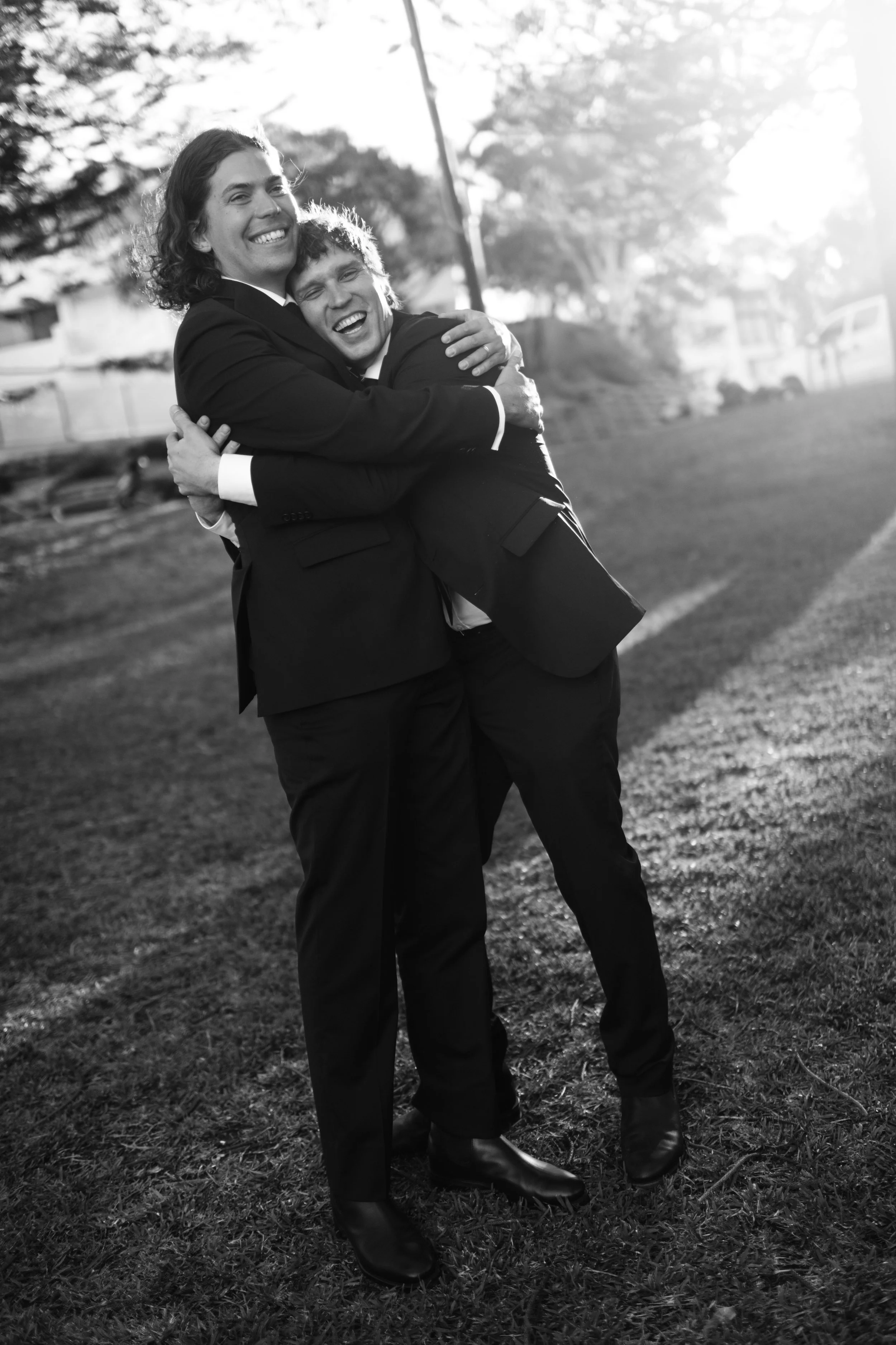 Two men in formal suits hugging outdoors in black and white, with trees and a park in the background, sunlight behind them.