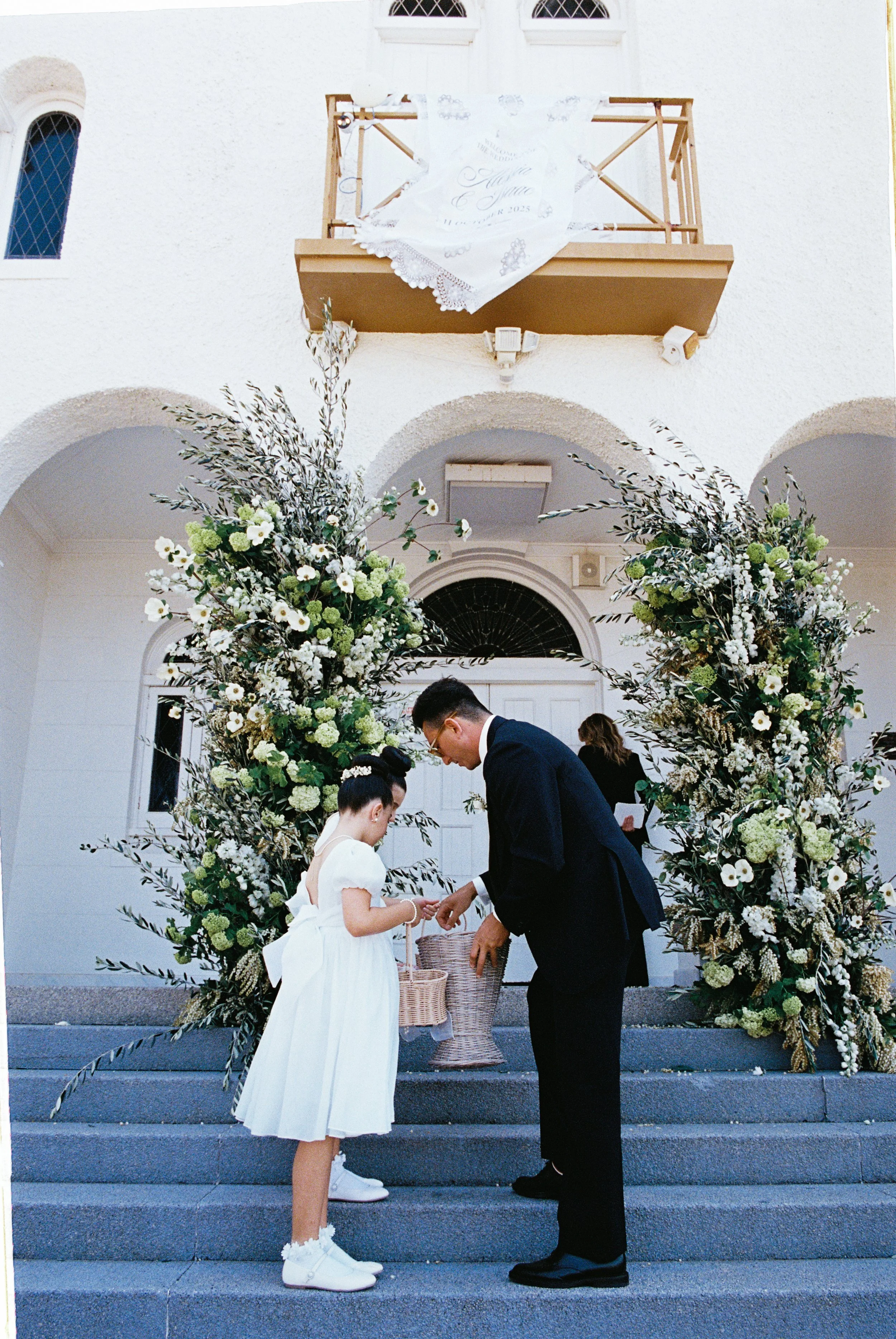 A wedding ceremony taking place on steps outside a white building with arched doorways, floral decorations, and a girl in a white dress and a man in a dark suit exchanging rings under a floral archway.
