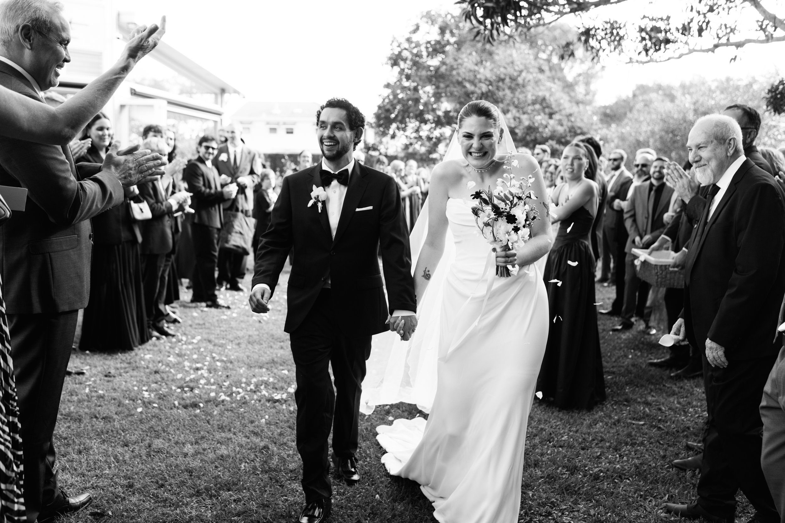 Black and white photo of a wedding reception outdoors with a happy bride and groom walking hand-in-hand amidst wedding guests.