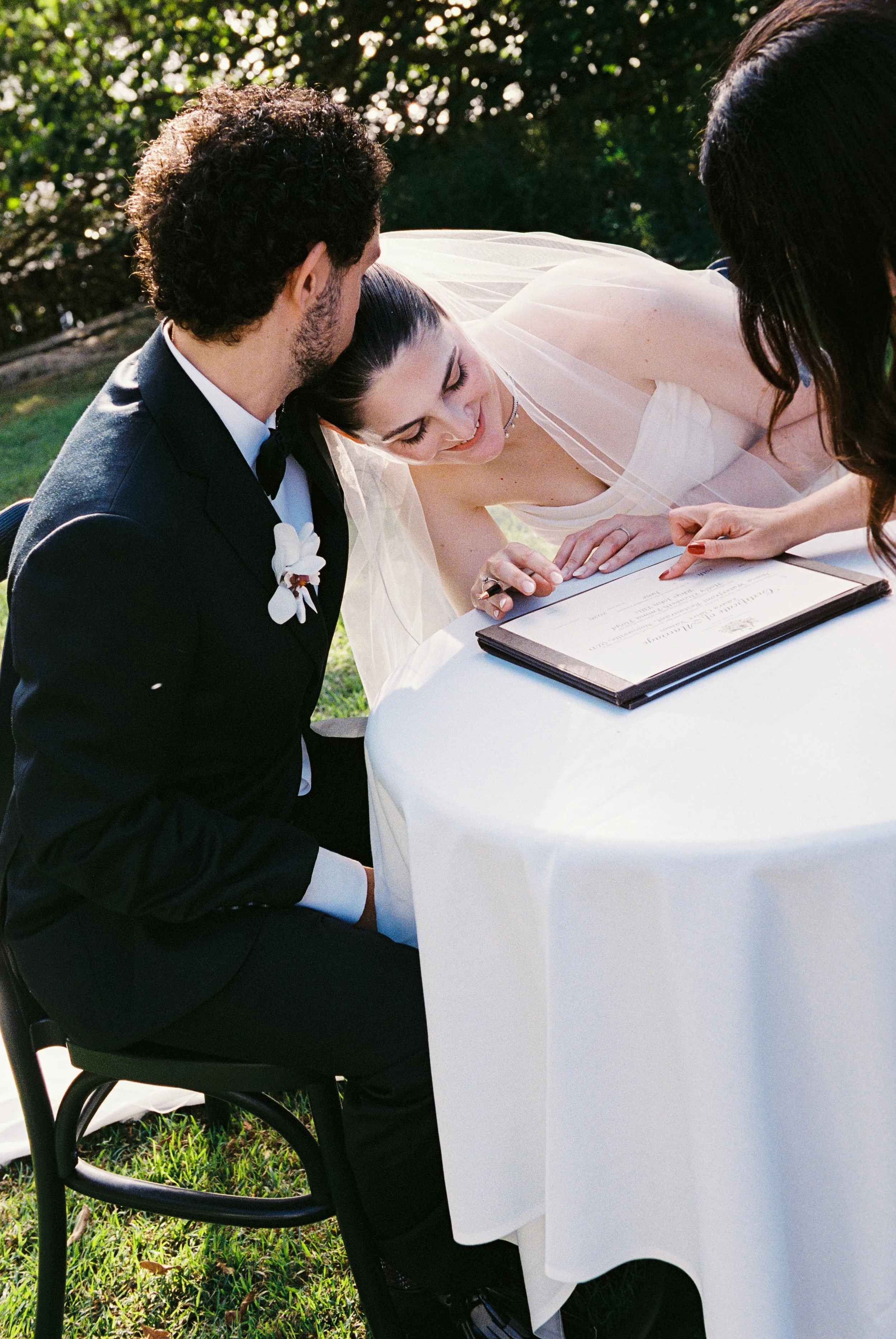 A bride and groom signing a marriage certificate outdoors at a white-clothed table, surrounded by greenery under sunlight.